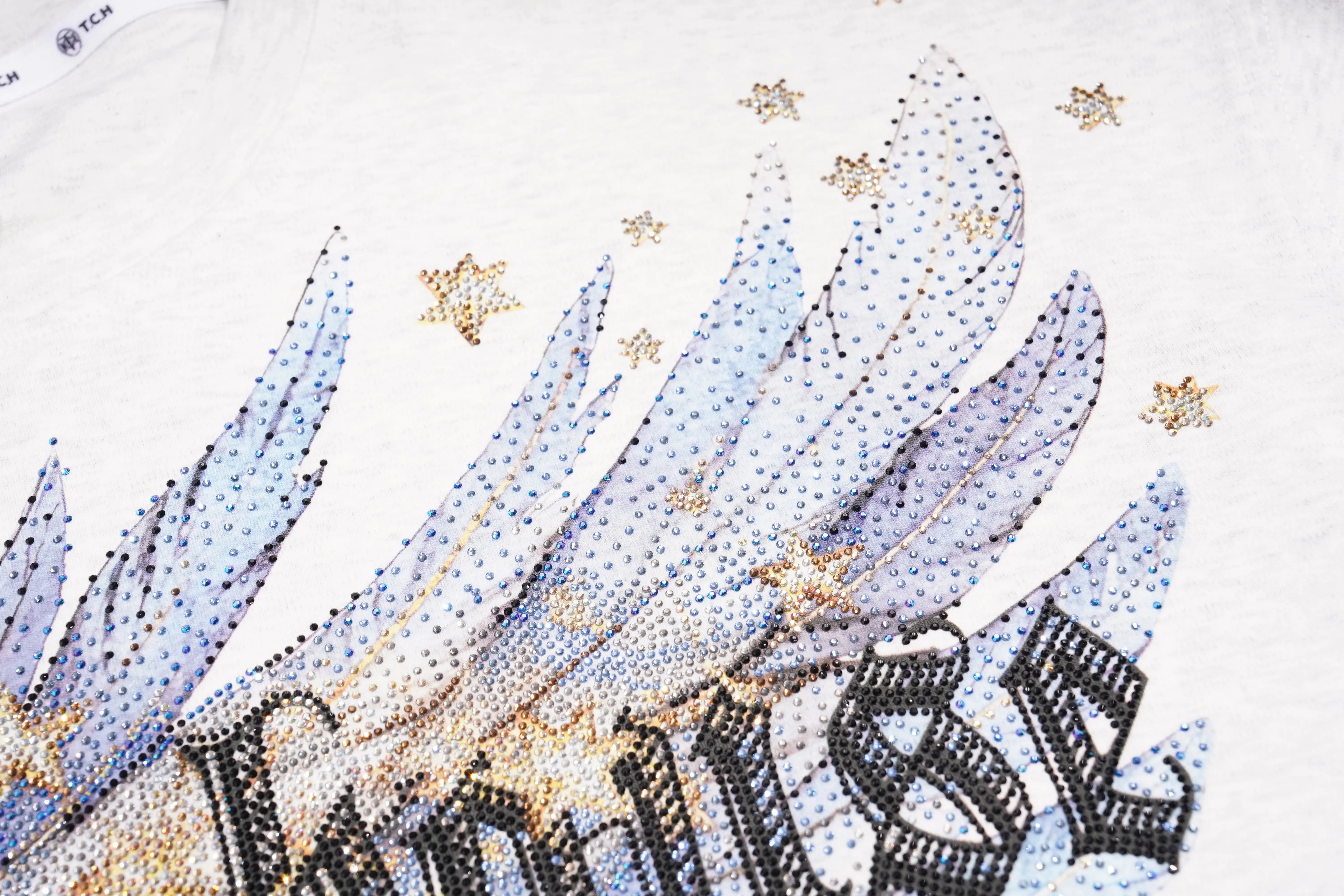 Close-up of a white Converse T-shirt featuring a rhinestone-embellished graphic of angel wings and the Converse script logo, accented with gold rhinestone stars. TCH Techno House Wing Graphic Oversized T-Shirt