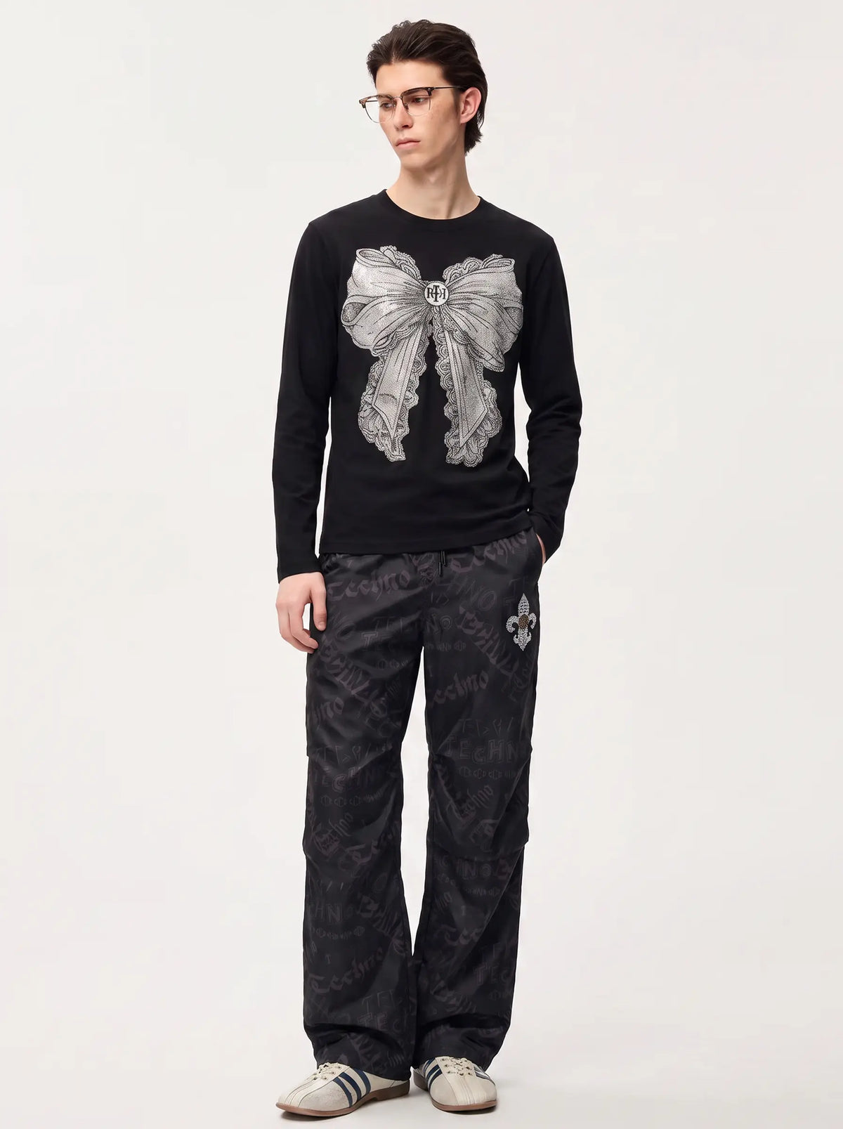 A man wearing a black long-sleeve crewneck sweater with a large gray bow graphic on the front, paired with dark patterned pants and light gray sneakers, standing against a plain light gray studio background. TCH Techno Logo Wide-Leg Cargo Pants