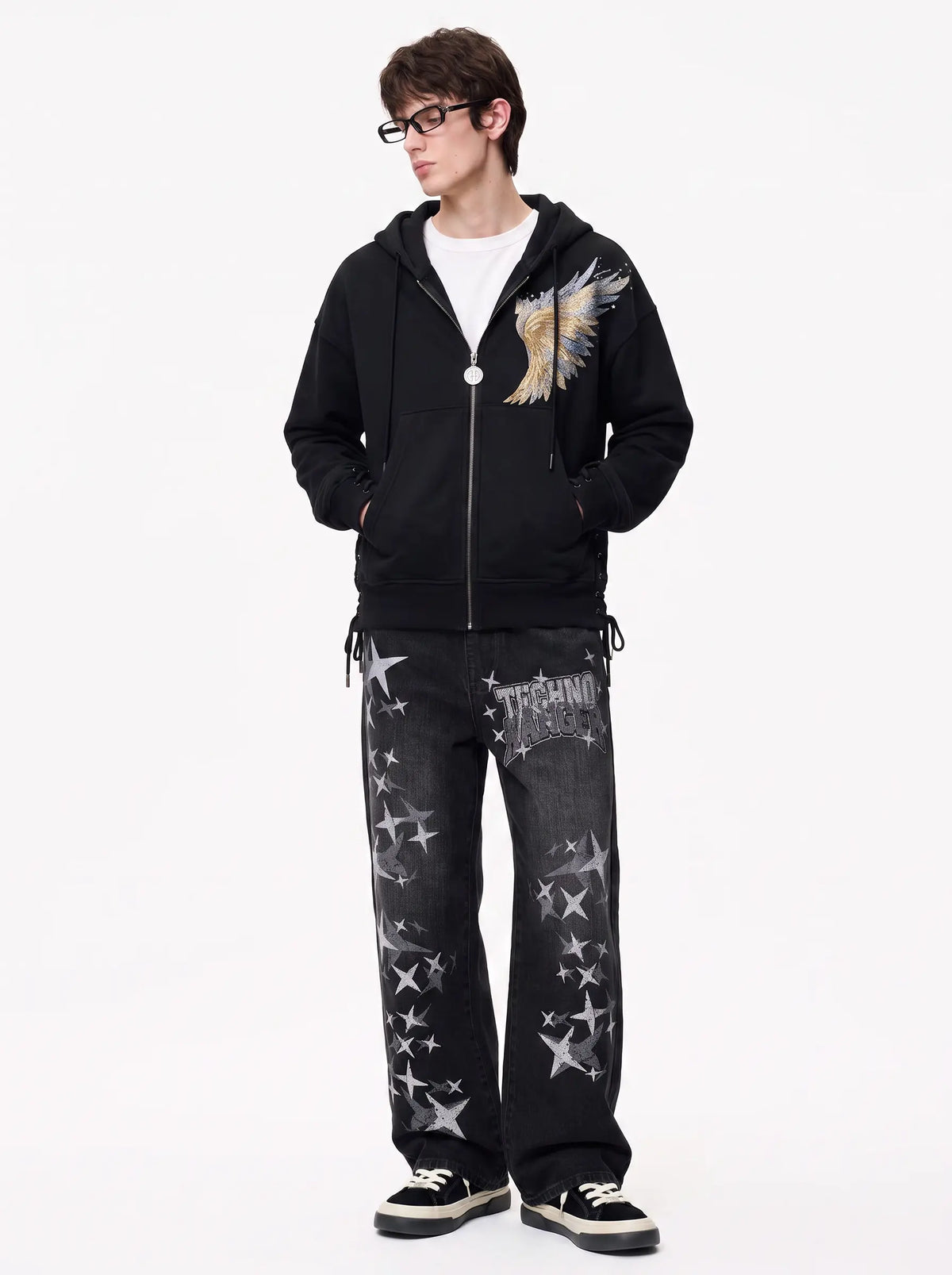 A model wearing a black zip-up hoodie with a large embroidered angel wing graphic on the chest, paired with black jeans featuring a distressed silver star pattern, a white t-shirt, and black sneakers. TCH Techno Magic Star Graphic Wide-Leg Jeans