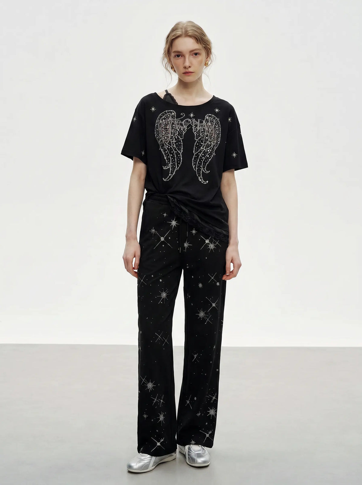 A model stands in a studio wearing a black short-sleeve T-shirt with a large silver wing graphic and matching black pants covered in small silver star-like sparkles, paired with silver sneakers. TCH Techno Rhinestone Angel Wings Lace Trim Off-Shoulder Top