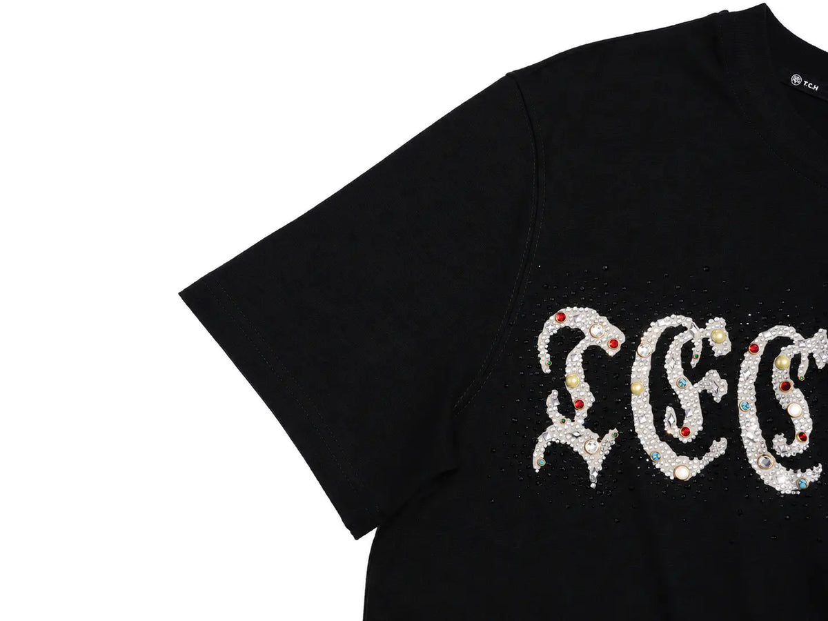 A close-up of a black short-sleeve T-shirt featuring a large decorative white gothic-style lettering graphic embellished with sequins and small red accents, shown against a plain white background. TCH Techno Rhinestone Gothic Graphic Black T-Shirt