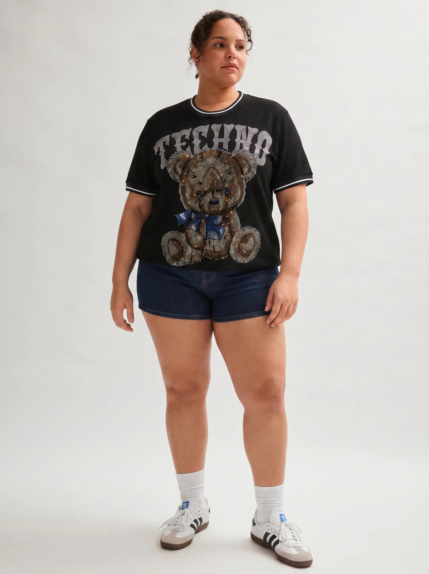 Person wearing a black t-shirt with a teddy bear graphic and 'Techno' text, dark shorts, and white sneakers on a white background. TCH Techno Teddy Rhinestone T-Shirt