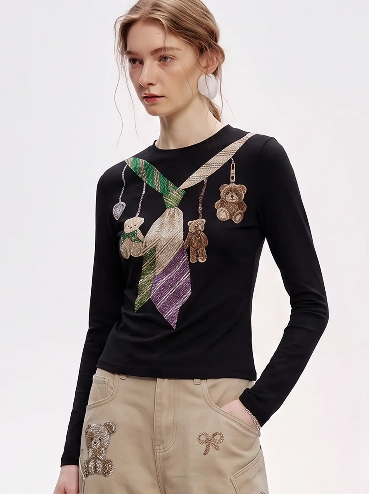 A model wearing a light blue denim jacket with various embroidered patches, including a teddy bear on the left chest and cherries on the sleeve, styled over a white t-shirt and dark pants. TCH Teddy Bear Charm Tie Graphic Black Long Sleeve T-Shirt