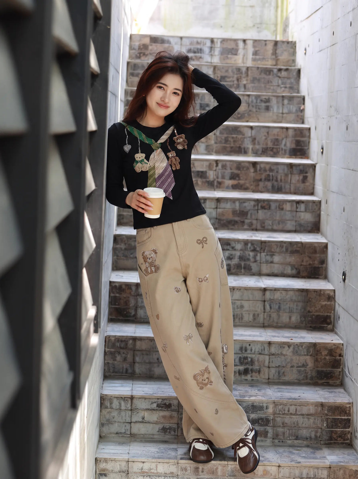 A woman posing on outdoor stone stairs wearing a black long-sleeve graphic top and beige wide-leg pants with teddy bear prints, holding a paper coffee cup. TCH Teddy Bear Charm Tie Graphic Black Long Sleeve T-Shirt