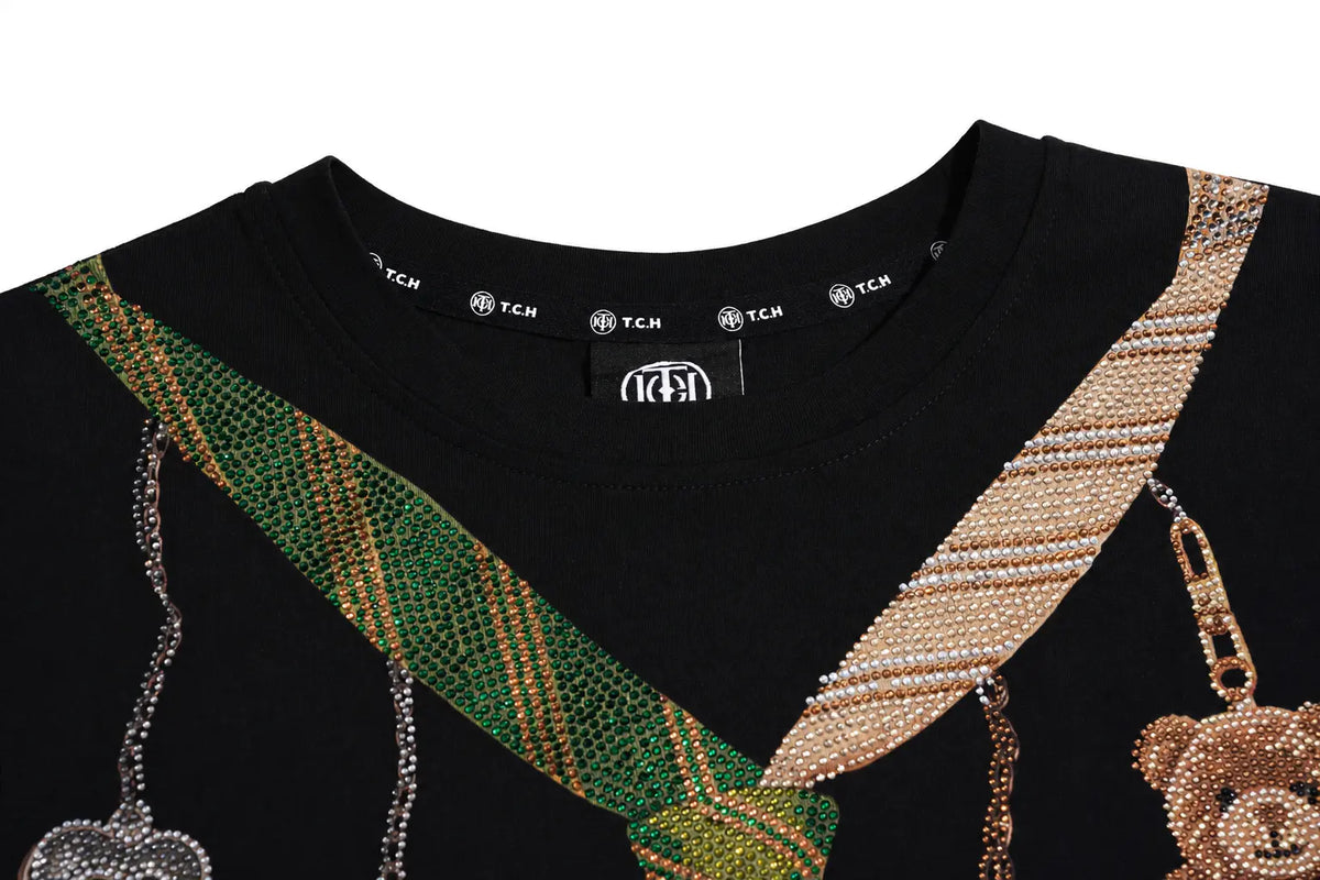 Close-up of a black TCH crewneck sweatshirt featuring a green and gold beaded tie design and a beige and gold beaded shoulder strap with a teddy bear motif. TCH Teddy Bear Charm Tie Graphic Black Long Sleeve T-Shirt