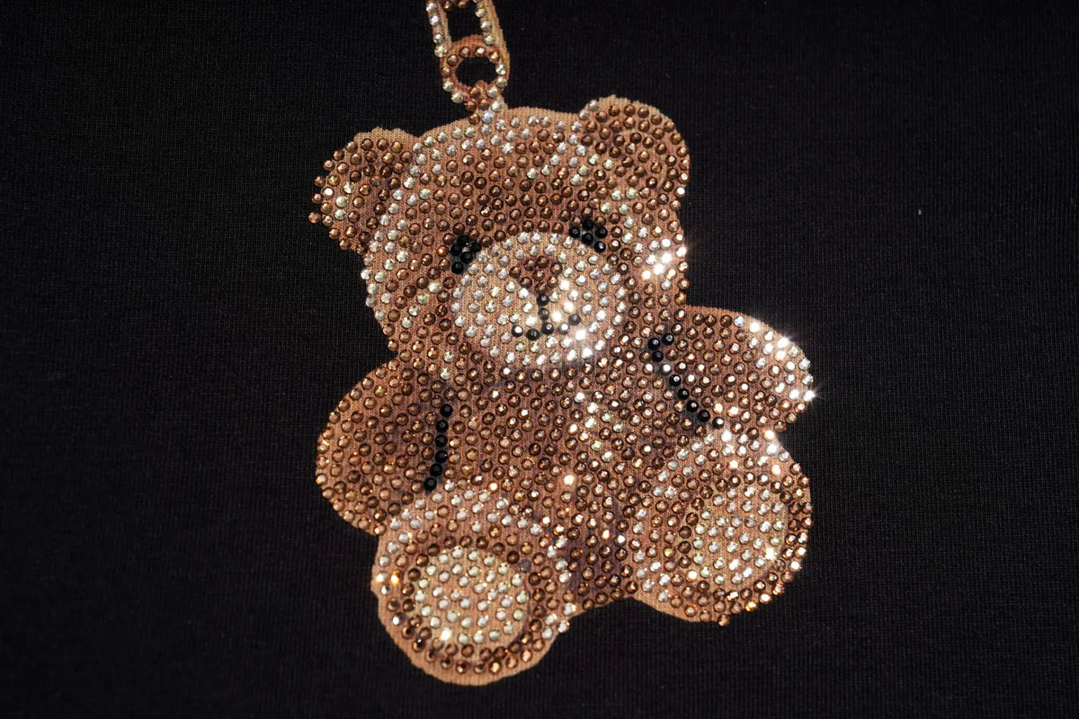 A teddy bear-shaped rhinestone keychain or bag charm in a rose gold tone, set against a solid black background. TCH Teddy Bear Charm Tie Graphic Black Long Sleeve T-Shirt