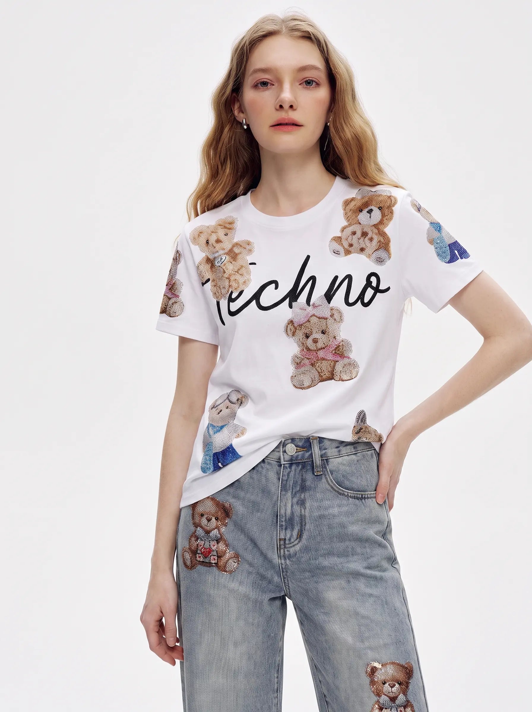 A woman modeling a white short-sleeve t-shirt with teddy bear prints and the word 'Techno' in black script across the chest, paired with light blue jeans featuring teddy bear patches. TCH Teddy Bear Rhinestone All-Over Graphic T-Shirt
