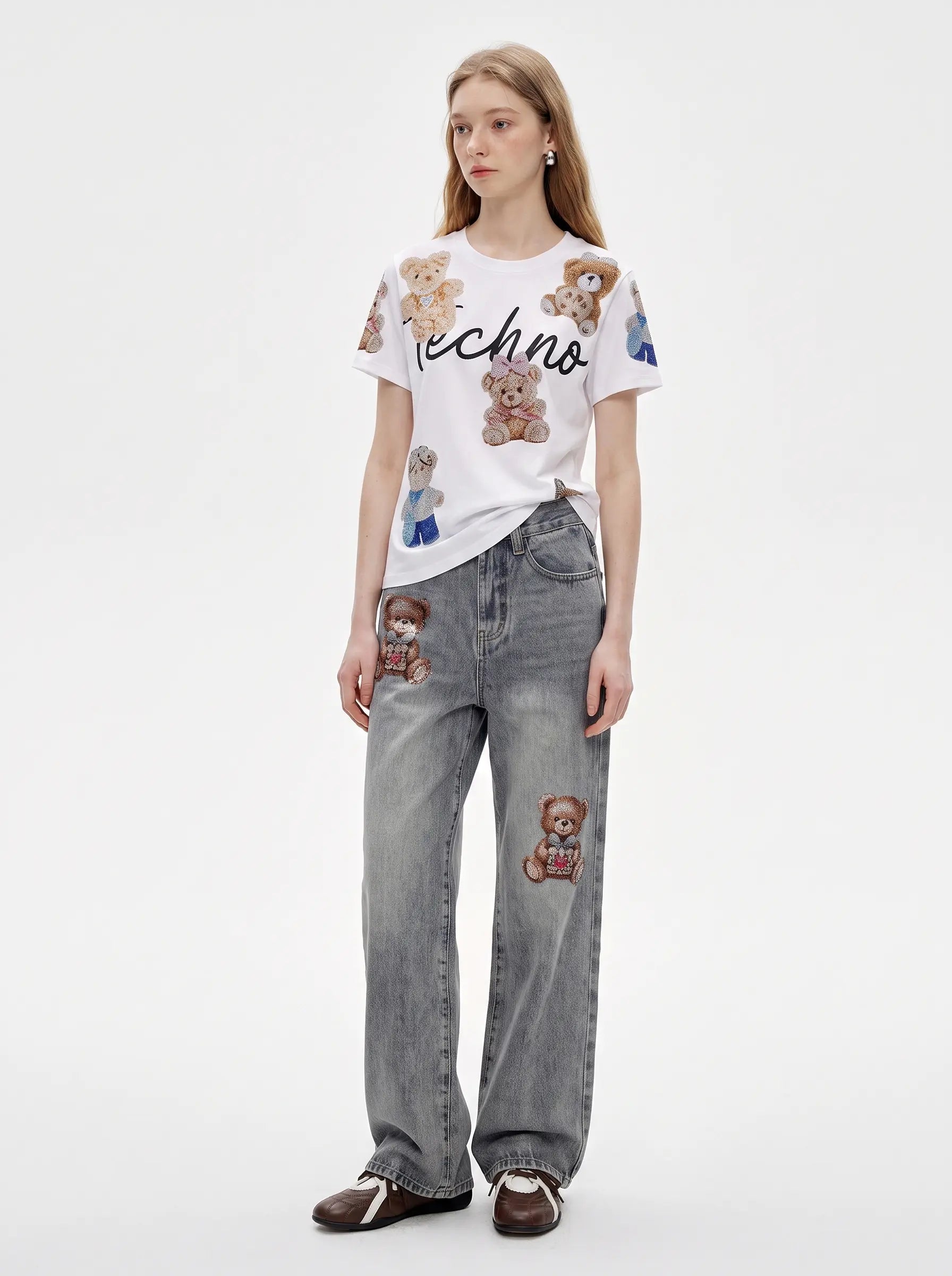 A model stands against a light gray studio background wearing a white short-sleeve T-shirt with teddy bear patches and the word 'techno' across the chest, paired with wide-leg gray jeans featuring teddy bear patches and dark brown loafers. TCH Teddy Bear Rhinestone All-Over Graphic T-Shirt