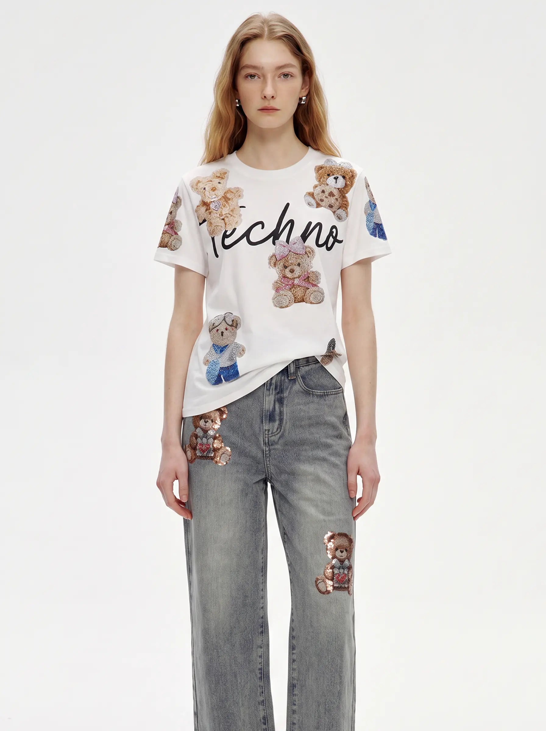 A model wearing a white short-sleeve t-shirt with teddy bear prints and the word 'techno' in black script, paired with gray jeans featuring teddy bear patches, against a plain white studio background. TCH Teddy Bear Rhinestone All-Over Graphic T-Shirt