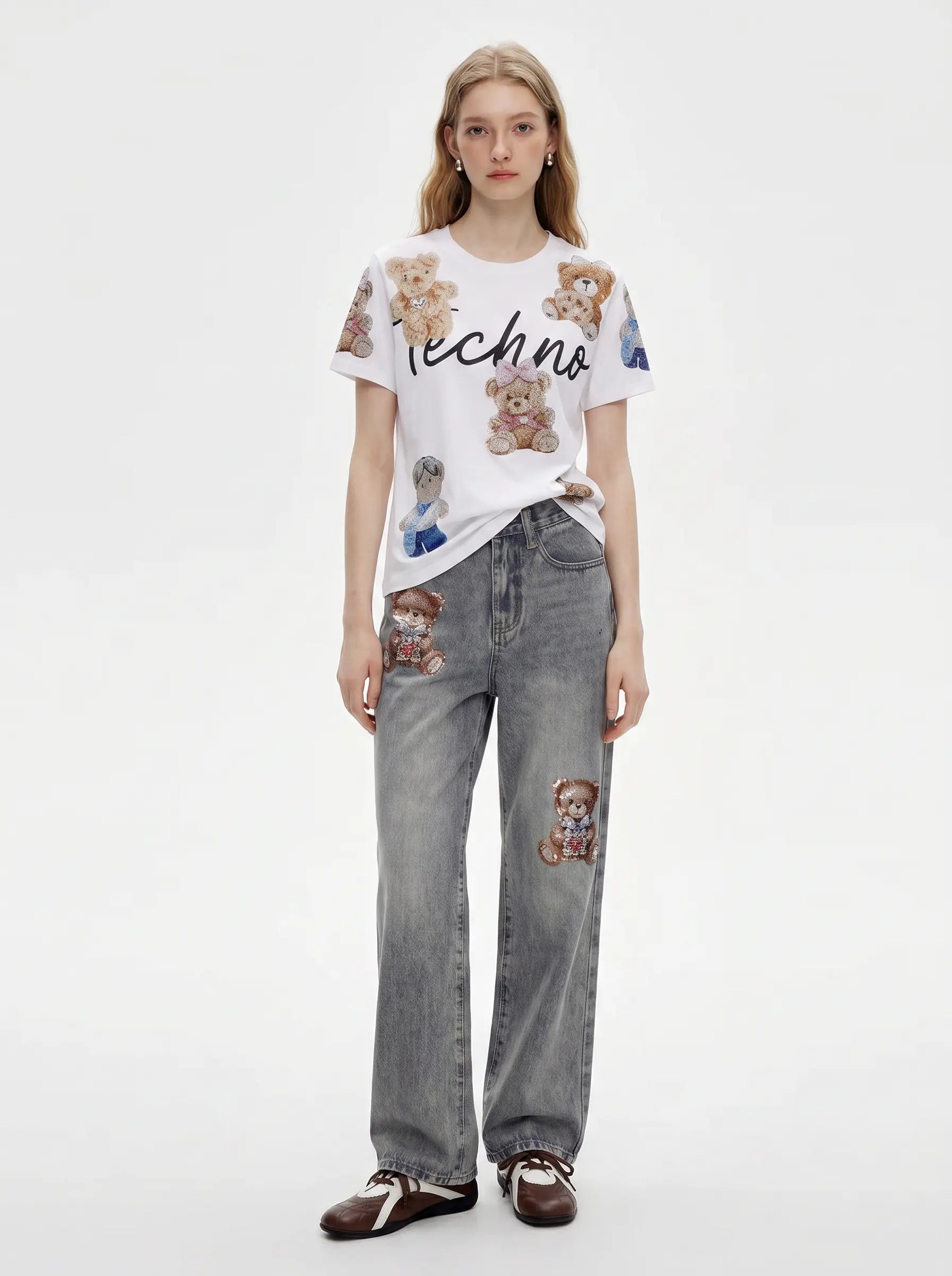 A model stands against a plain light gray studio background wearing a white graphic T-shirt with teddy bear prints and the word 'techno' across the chest, paired with light gray jeans featuring teddy bear patches and dark brown loafers. TCH Teddy Bear Rhinestone All-Over Graphic T-Shirt