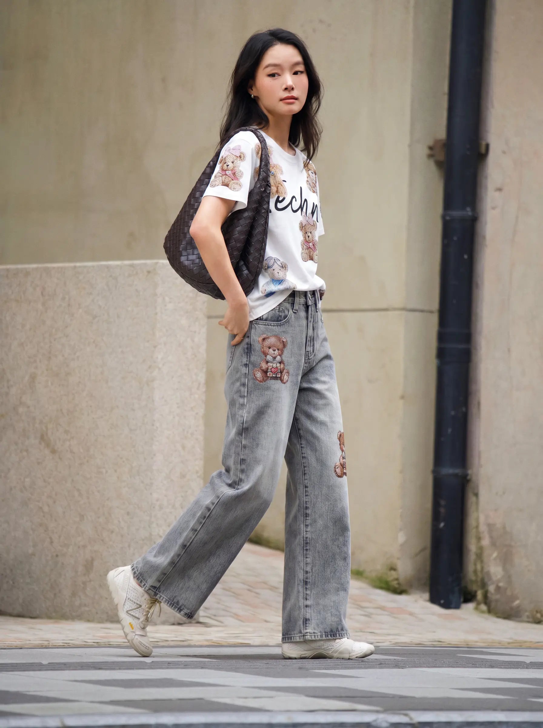 A woman walking outdoors wearing a white graphic T-shirt, light gray wide-leg jeans with teddy bear patches, white sneakers, and carrying a dark patterned shoulder bag. TCH Teddy Bear Rhinestone All-Over Graphic T-Shirt