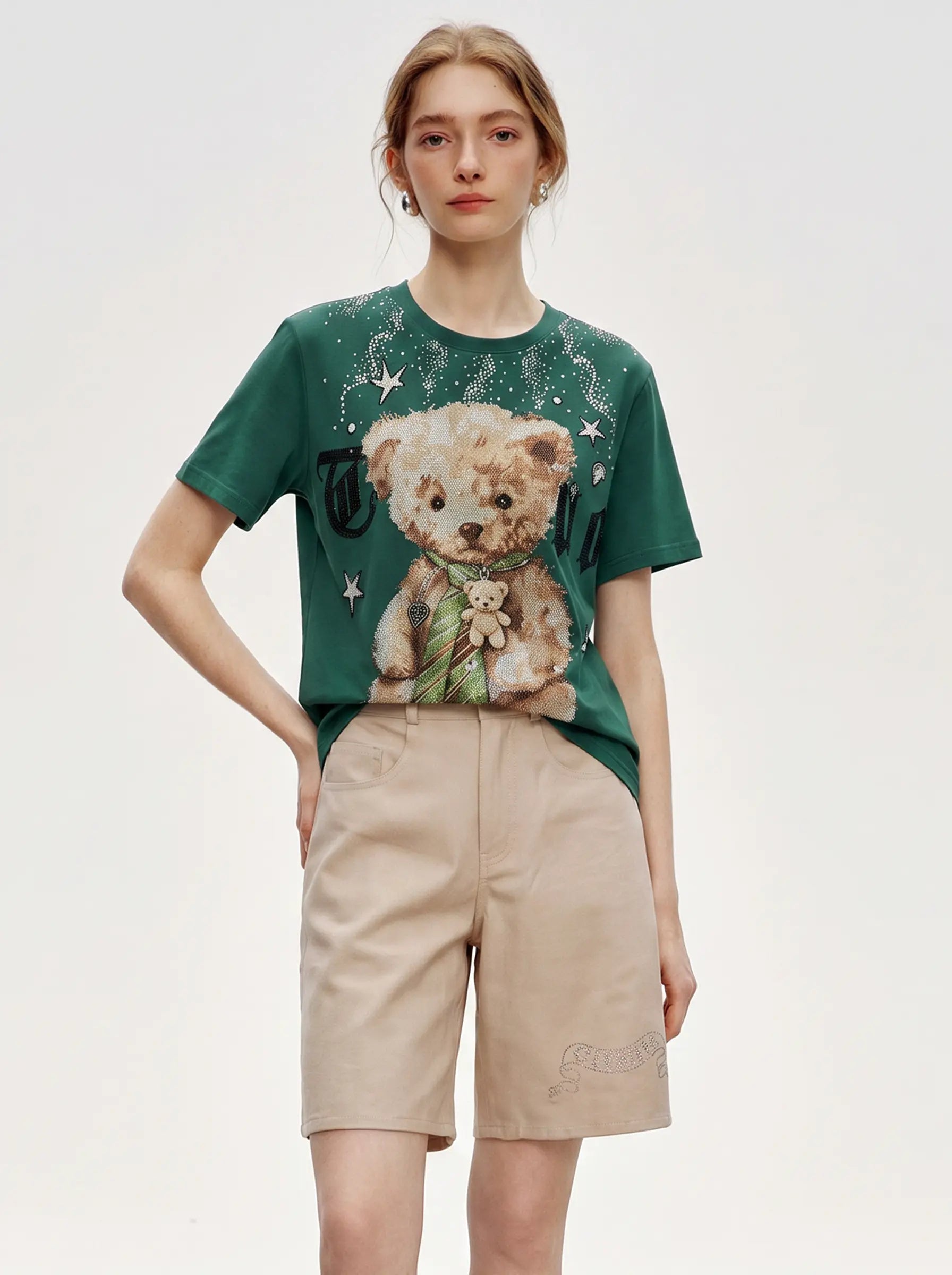 A model wearing a green graphic t-shirt with a teddy bear print and beige shorts, standing against a plain light gray studio background. TCH Teddy Bear Rhinestone Graphic T-Shirt