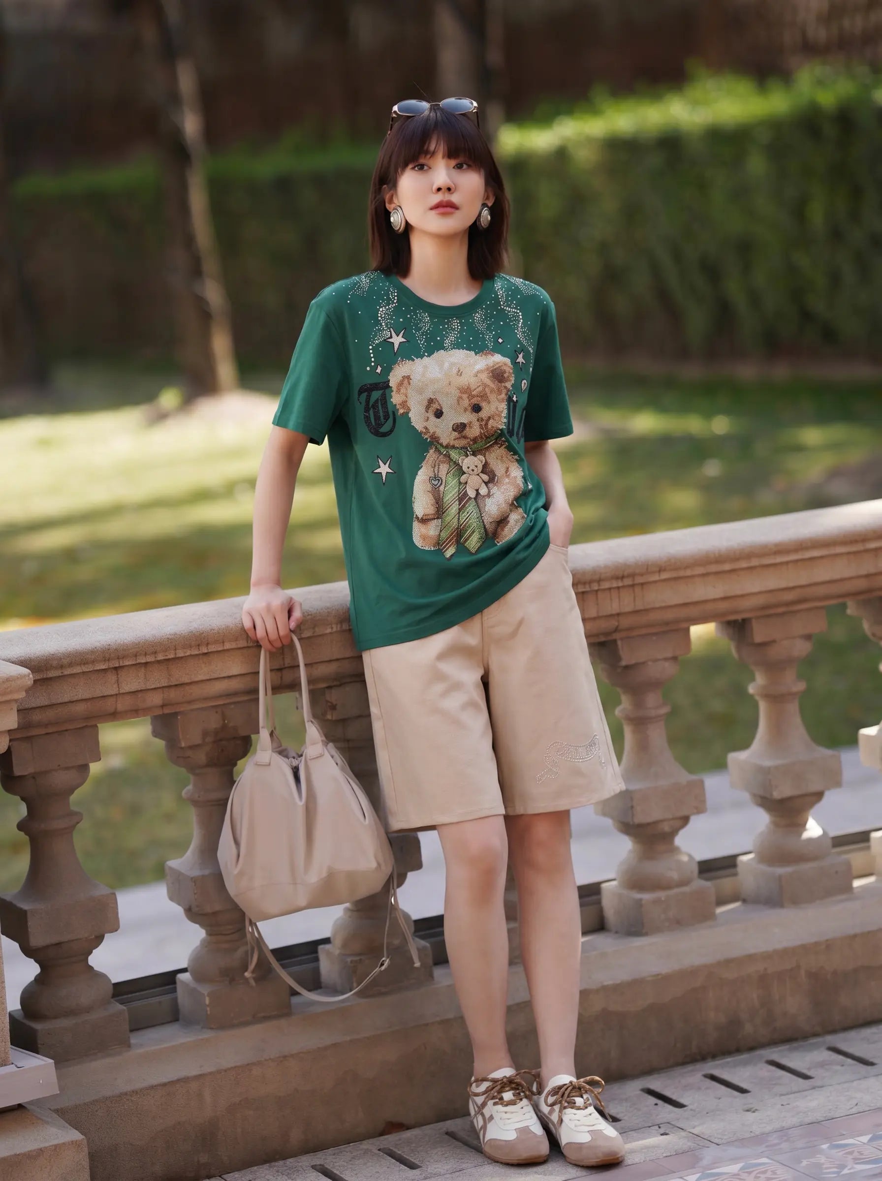 A woman stands outdoors on a stone bridge wearing a green graphic T-shirt with a teddy bear print, beige shorts, and white sneakers, while holding a beige handbag. TCH Teddy Bear Rhinestone Graphic T-Shirt