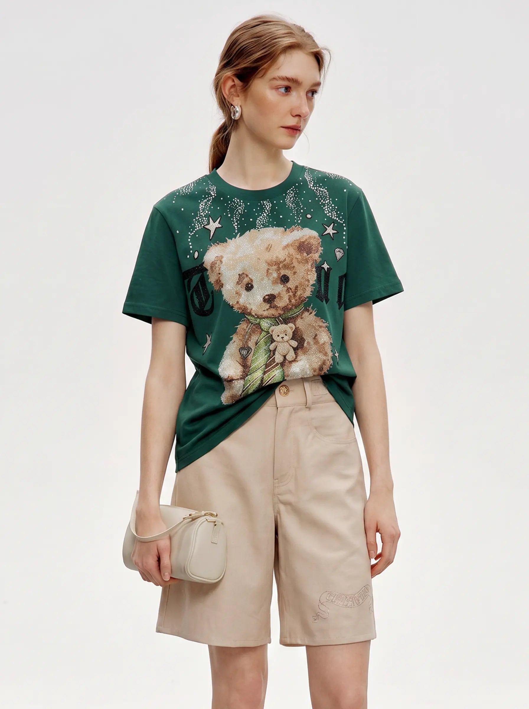 A woman modeling a dark green graphic t-shirt with a teddy bear print, paired with beige shorts and holding a small beige handbag against a light gray studio background. TCH Teddy Bear Rhinestone Graphic T-Shirt