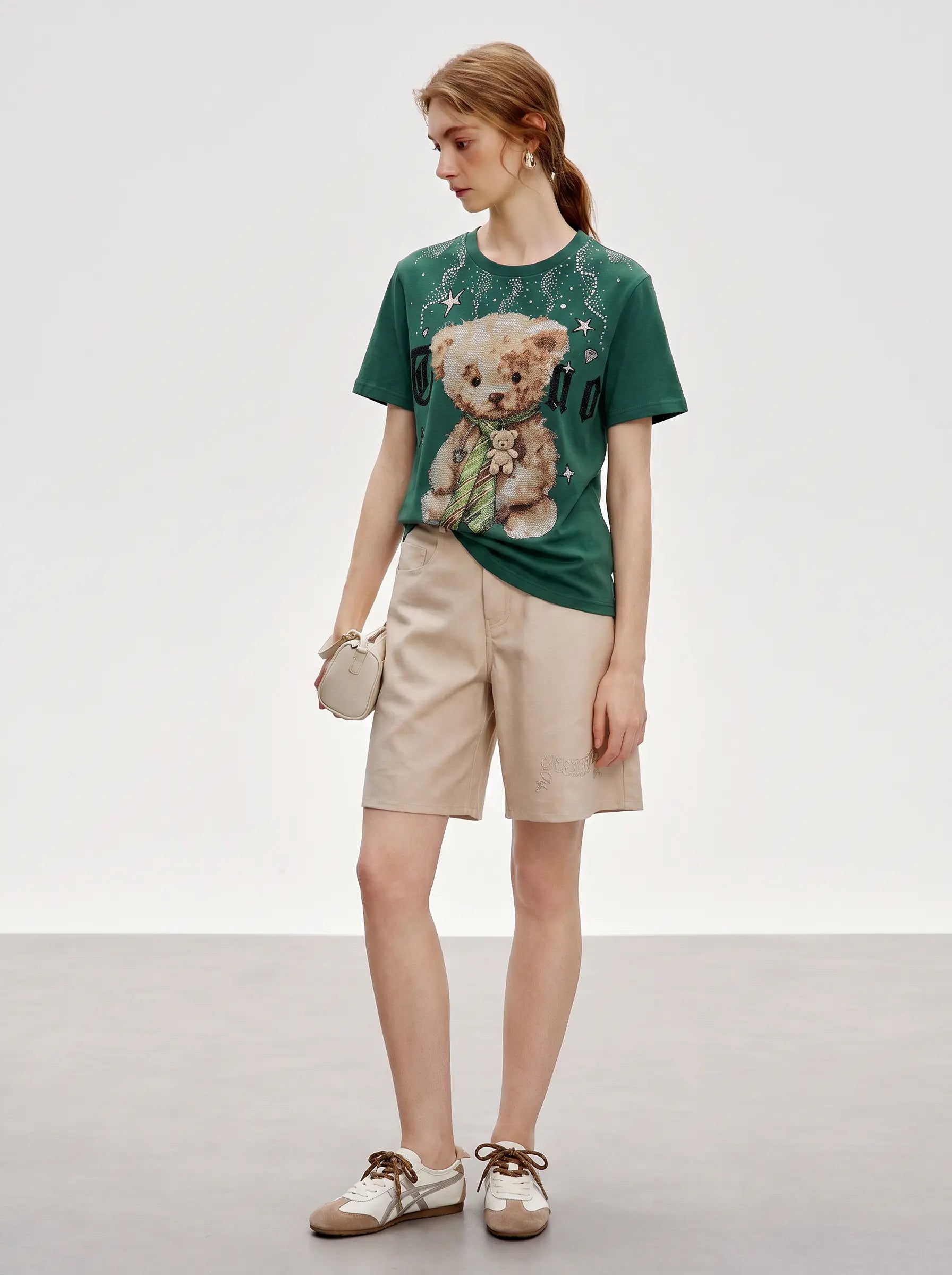 A model stands in a studio wearing a green graphic T-shirt with a teddy bear print, beige shorts, and white-and-brown shoes, holding a small beige handbag. TCH Teddy Bear Rhinestone Graphic T-Shirt