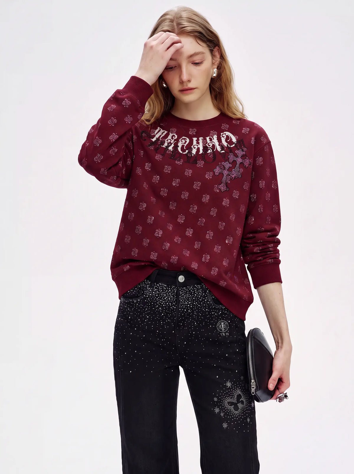 A model wearing a burgundy long-sleeve sweatshirt with a repeating monogram pattern and 'MACHNO' text across the chest, paired with black embellished pants and holding a black clutch against a plain white background. TCH Vintage Paisley Graphic Burgundy Crewneck Sweatshirt