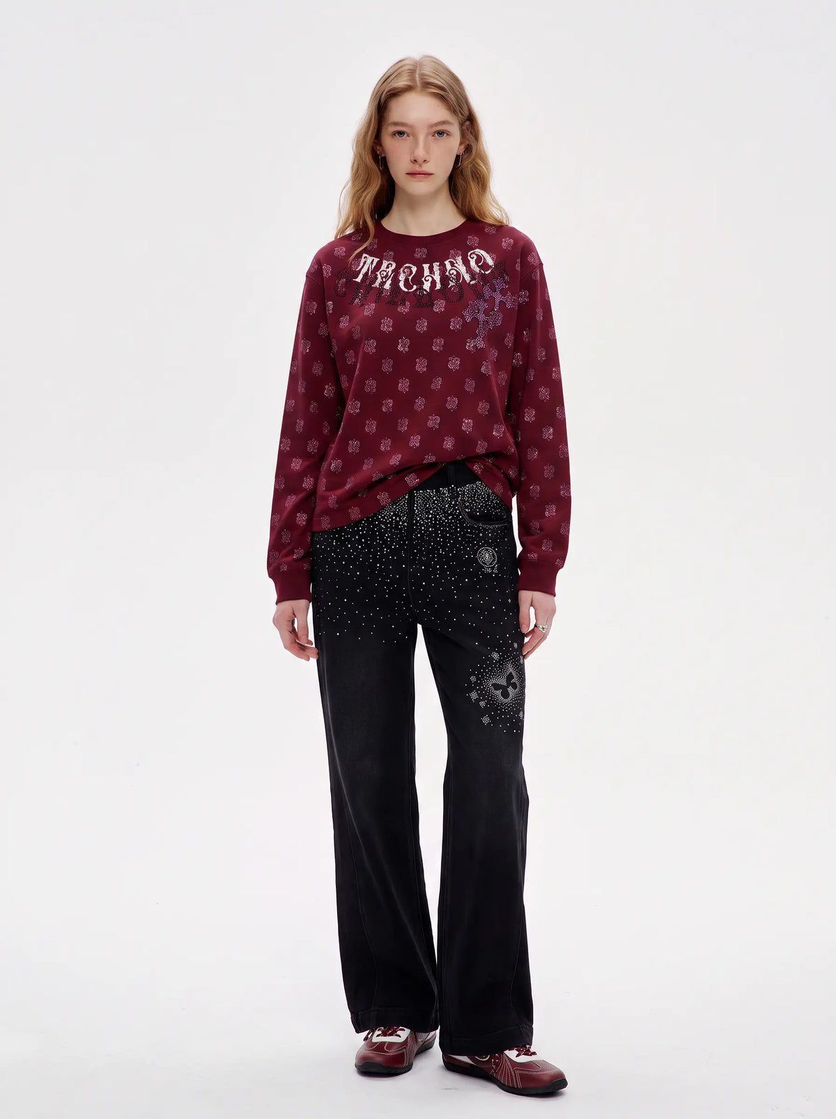 A model stands against a plain white studio background wearing a burgundy crewneck sweatshirt with a repeating monogram pattern and the word 'PACHIO' across the chest, paired with black wide-leg pants and burgundy loafers. TCH Vintage Paisley Graphic Burgundy Crewneck Sweatshirt