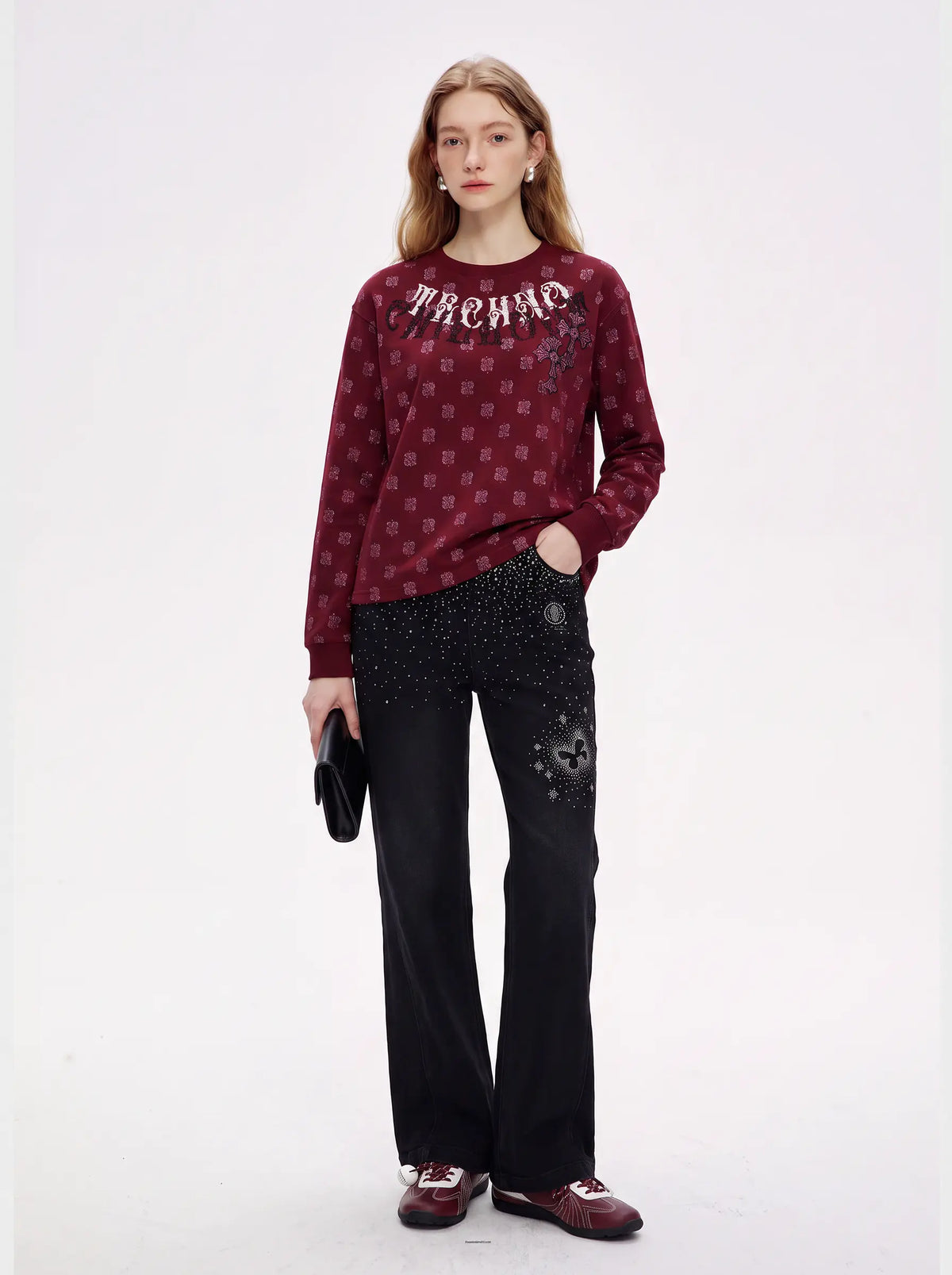 A model stands against a plain white studio background wearing a burgundy long-sleeve knit top with a repeating small pattern and 'MACHADO' lettering across the chest, paired with black pants featuring silver embellishments and burgundy shoes. TCH Vintage Paisley Graphic Burgundy Crewneck Sweatshirt