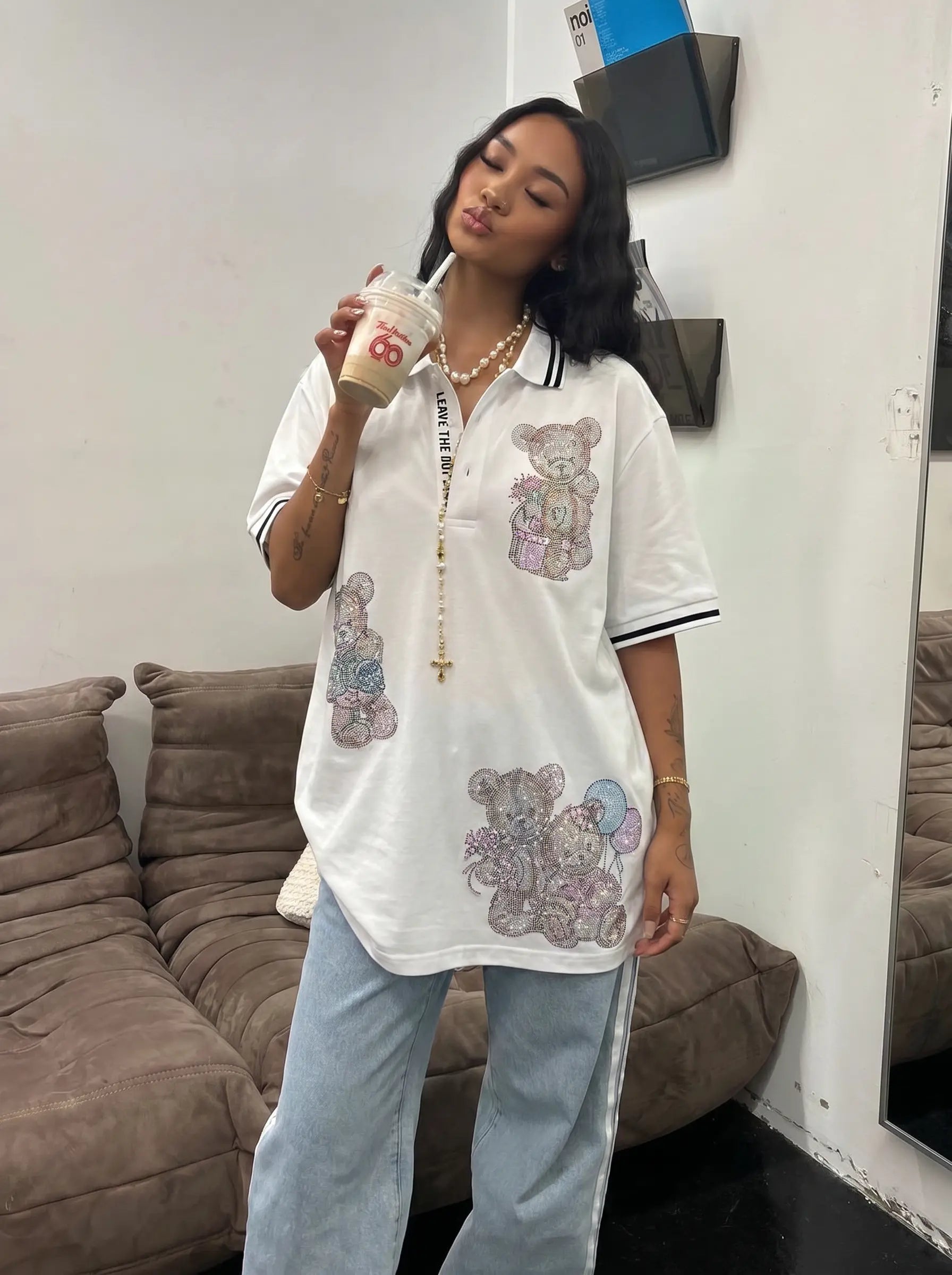 A woman wearing an oversized white short-sleeve polo shirt with teddy bear graphics and light blue jeans, holding an iced coffee in a living room setting. TCH White Teddy Bear Embellished Polo Shirt