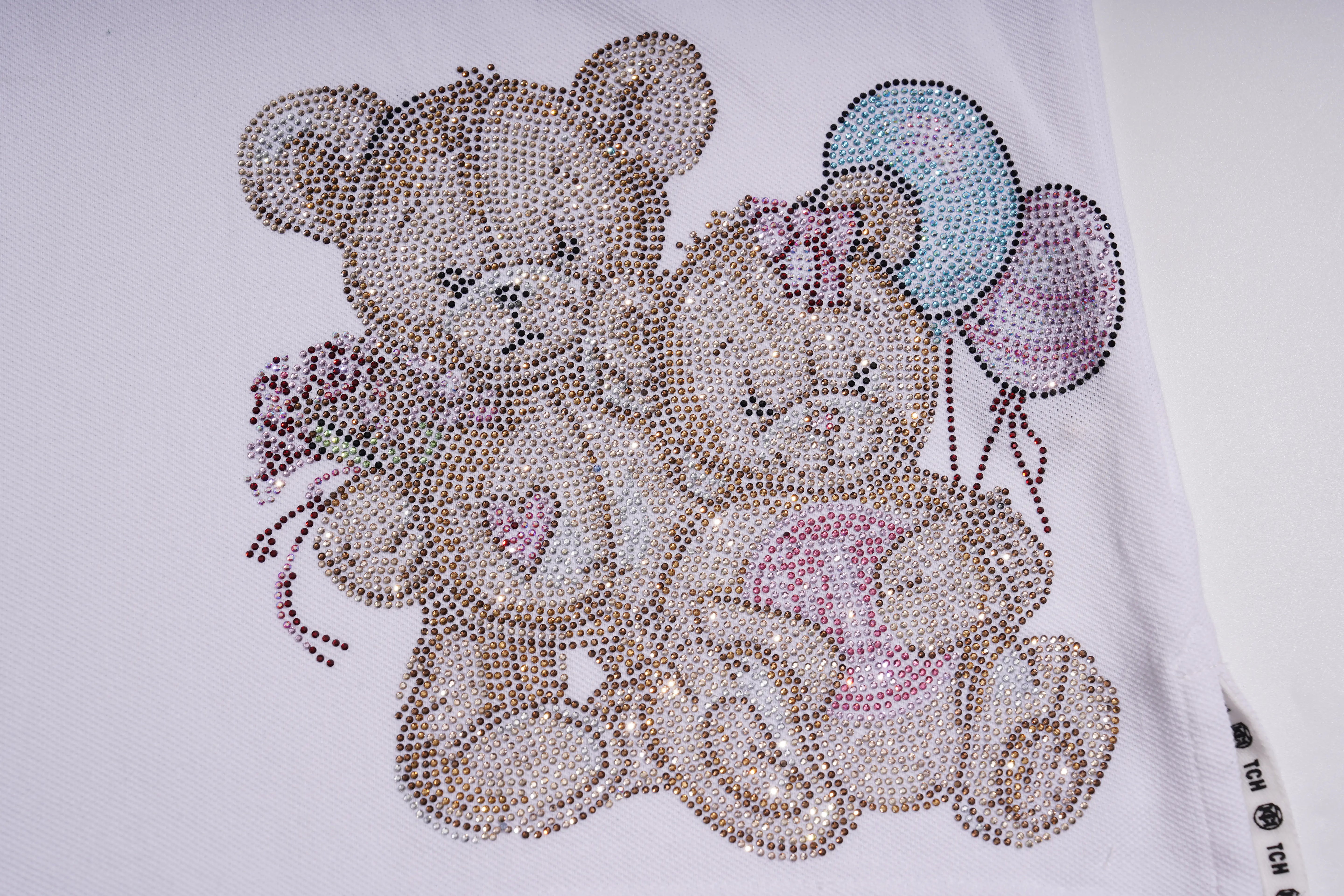 A close-up of a white garment featuring a rhinestone-style teddy bear and baby design with pastel balloons. TCH White Teddy Bear Embellished Polo Shirt