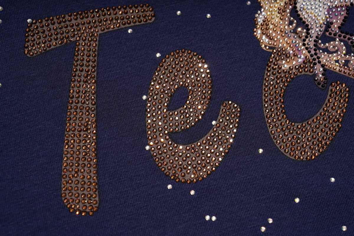 Close-up of a navy blue garment featuring large letter 'T' and 'e' embellished with brown rhinestones, with a matching rhinestone design partially visible on the right. TCH Winged Unicorn Starry Rhinestone Long Sleeve T-Shirt