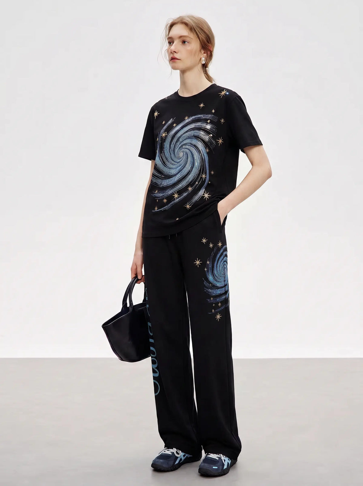 A woman modeling a black short-sleeve t-shirt and matching wide-leg pants set, both featuring a blue spiral galaxy print with small gold star details. She is holding a black handbag and wearing dark blue sneakers, set against a plain light gray studio background. TCH Women’s Black Wide-Leg Sweatpants with Rhinestone Star Graphic