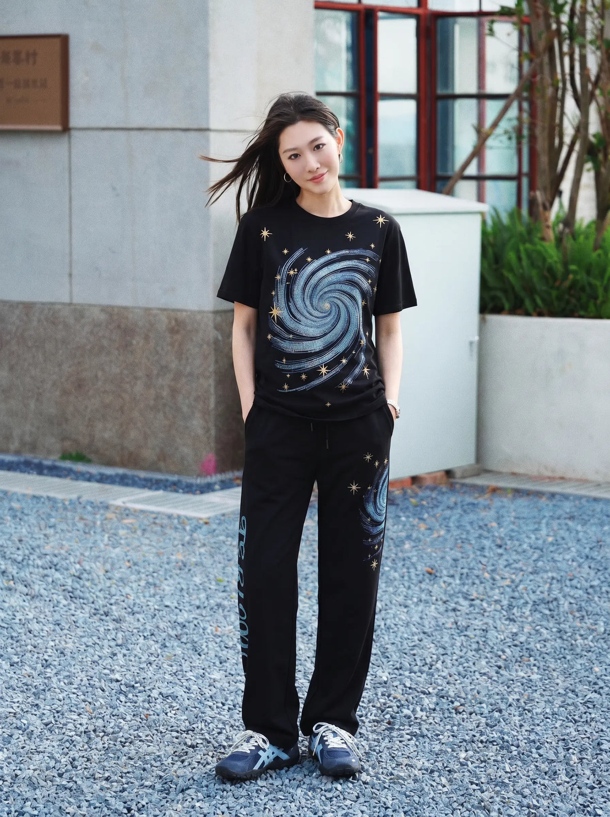 A woman stands outdoors on a gravel surface wearing a matching black two-piece set with a galaxy swirl print on the t-shirt and pants, paired with blue sneakers. TCH Women’s Black Wide-Leg Sweatpants with Rhinestone Star Graphic