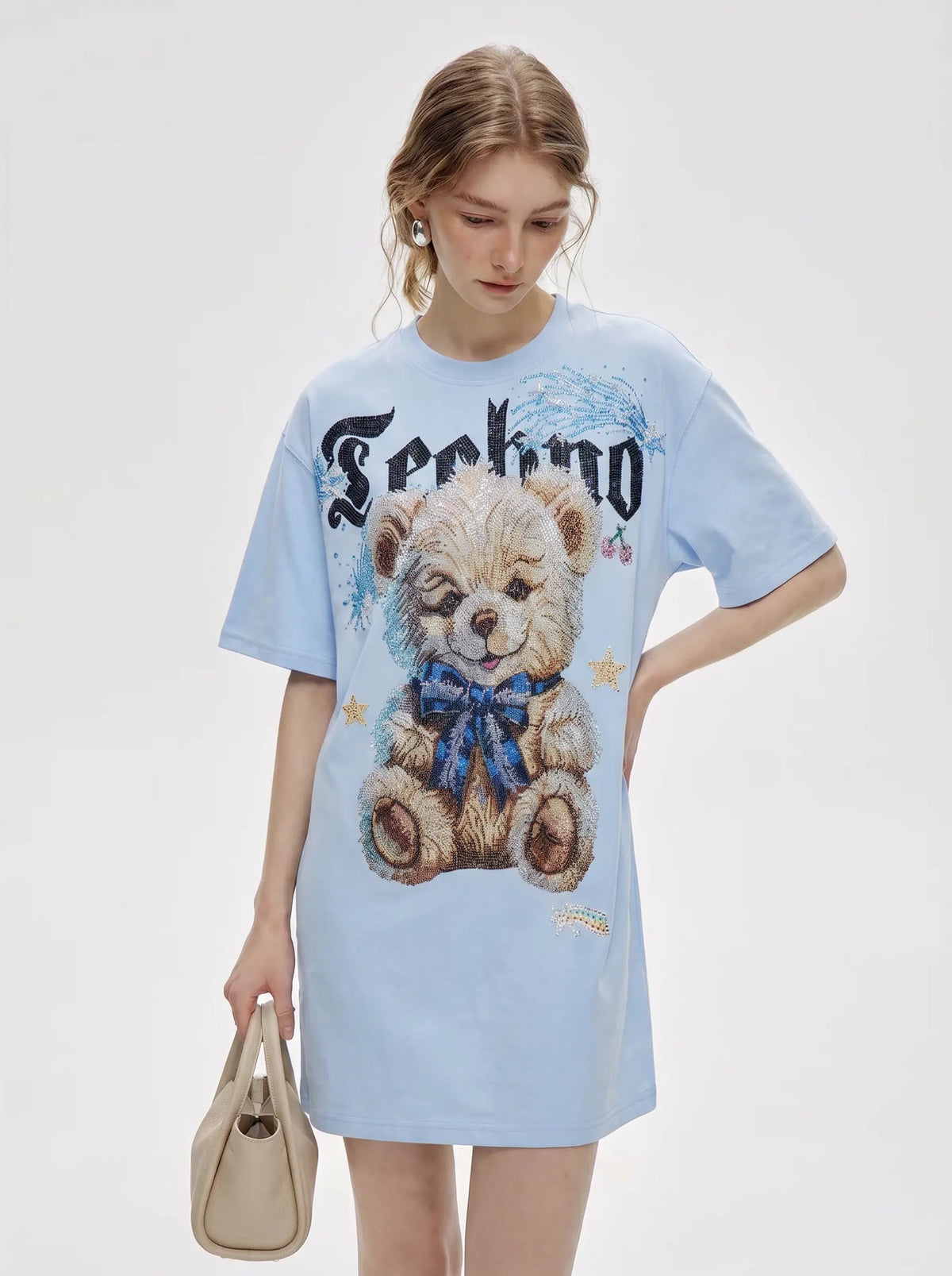 A woman modeling an oversized light blue t-shirt dress featuring a large teddy bear graphic and the word 'Techno' in a gothic font, holding a beige handbag against a plain light gray background. TCH Women's Blue Teddy Bear Graphic Oversized T-Shirt