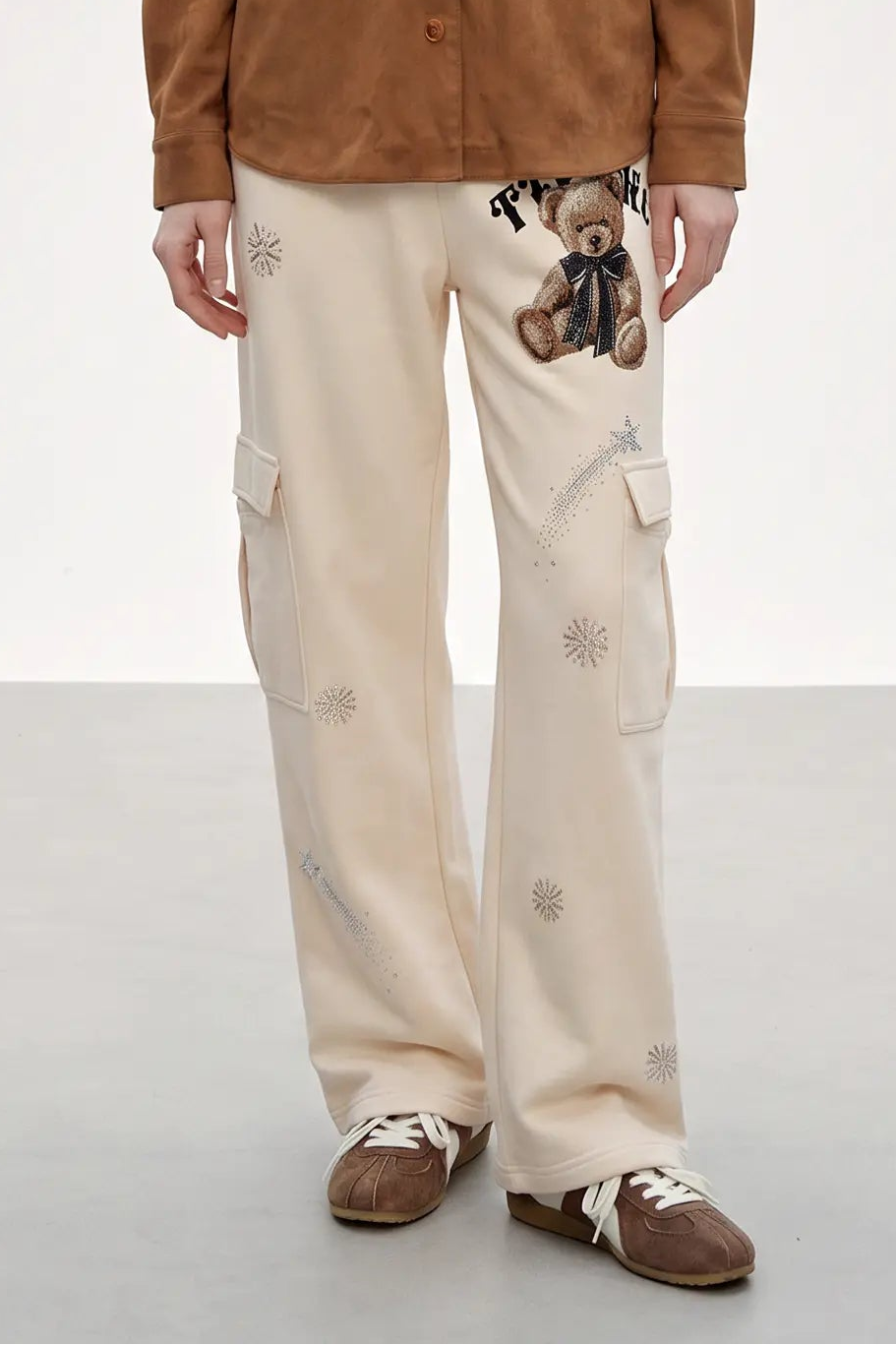 A person wearing cream cargo pants with a teddy bear graphic and embroidered star details, paired with a brown jacket and brown shoes, shown from the waist down against a light studio background. TCH Women’s Cream Cargo Sweatpants with Teddy Bear Rhinestone Graphic