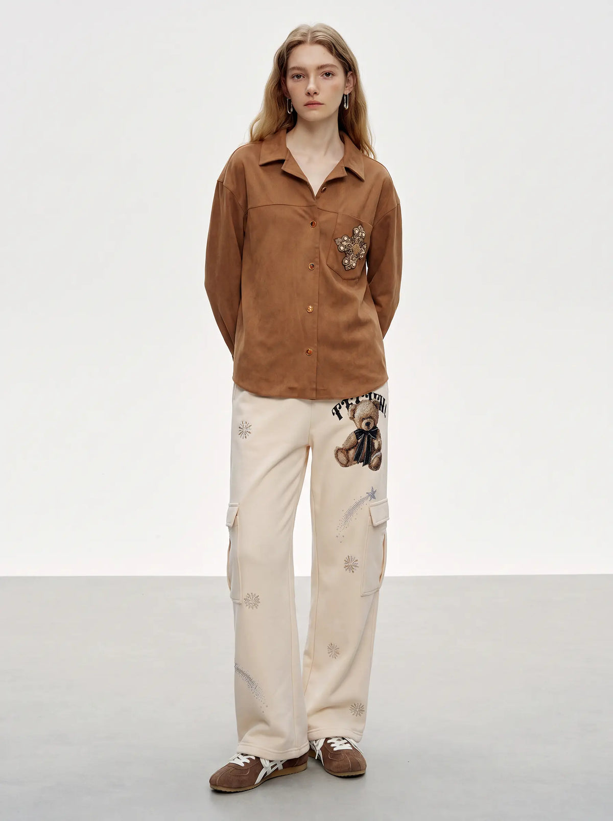 A model stands in a studio wearing a brown button-up shirt with floral embroidery on the chest pocket, paired with off-white cargo pants featuring teddy bear and floral embroidery, and brown slip-on shoes. TCH Women’s Cream Cargo Sweatpants with Teddy Bear Rhinestone Graphic