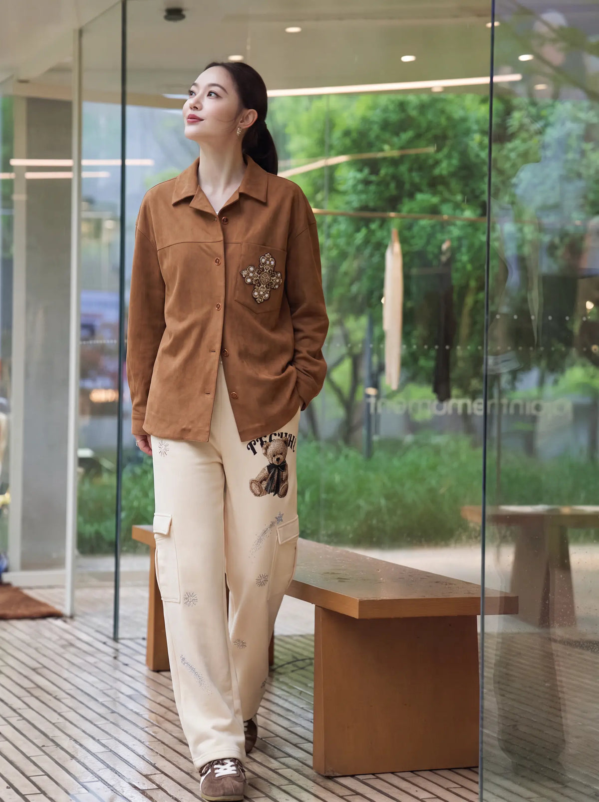 A woman stands in a modern glass-walled indoor space wearing a brown button-up shirt with a floral applique on the chest and off-white cargo pants with printed details. TCH Women’s Cream Cargo Sweatpants with Teddy Bear Rhinestone Graphic