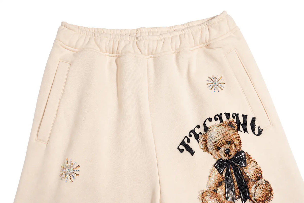 Cream-colored sweatpants featuring a teddy bear graphic with a black bow tie and the word 'TECHNIC' embroidered on the leg, along with rhinestone snowflake embellishments. TCH Women’s Cream Cargo Sweatpants with Teddy Bear Rhinestone Graphic