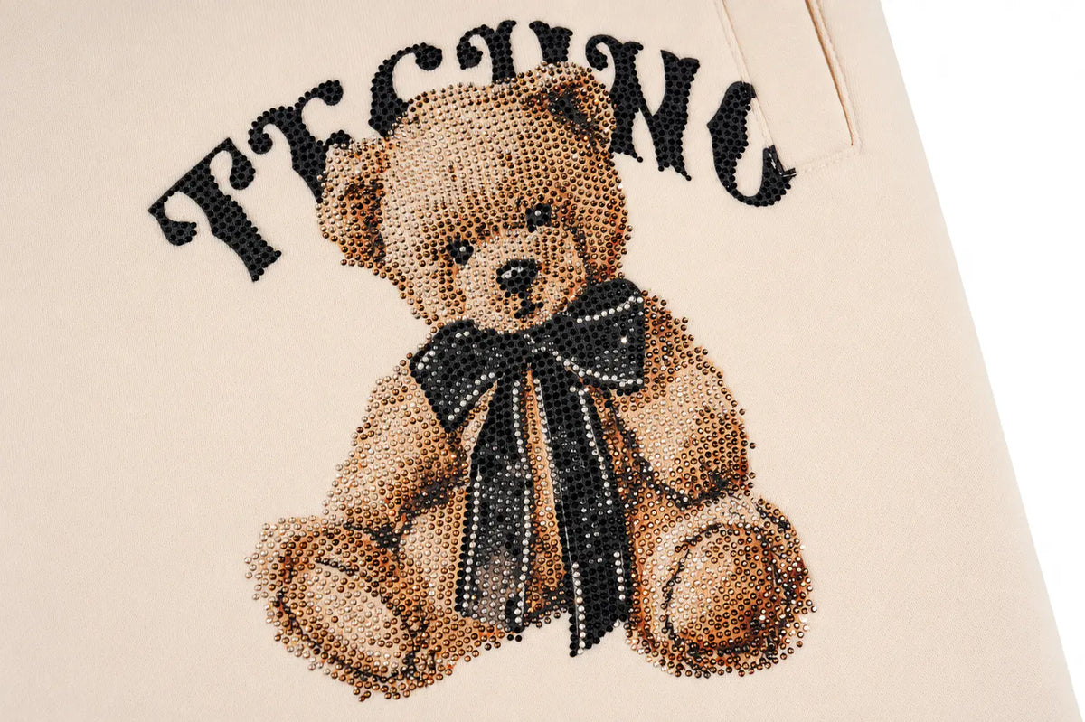 A close-up of a beige fabric surface featuring a teddy bear design made of rhinestones, with a black bow and the word 'TEACHING' in black above it. TCH Women’s Cream Cargo Sweatpants with Teddy Bear Rhinestone Graphic