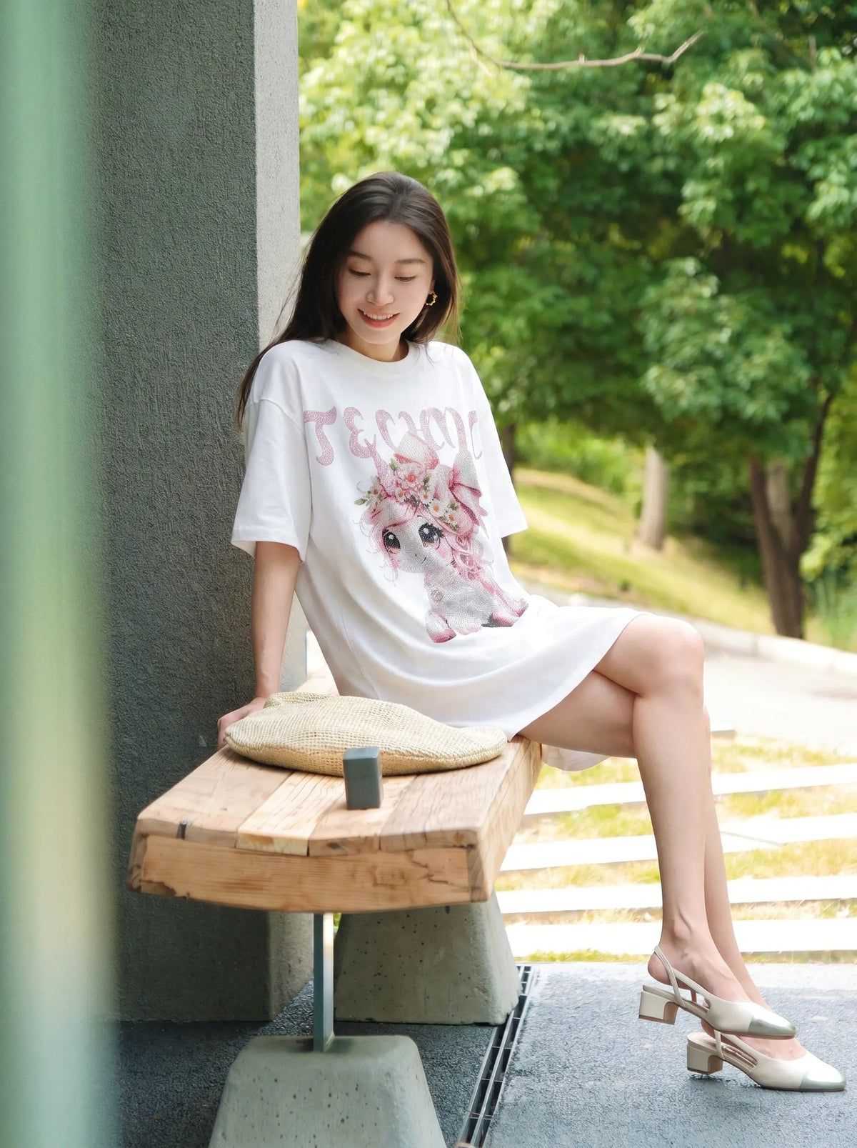A woman sits on a wooden bench outdoors wearing a white oversized graphic T-shirt dress with a pink floral print and the word 'FEMEN' in pink. She is wearing beige block-heel mule sandals, with a woven straw hat resting on the bench in front of her. TCH Women's Pink Angel Graphic Oversized T-Shirt
