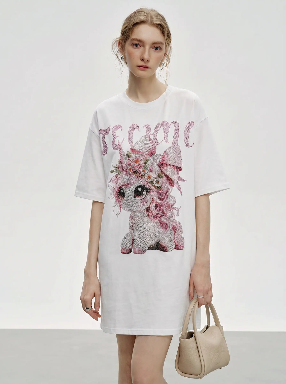A model wearing a white oversized T-shirt dress with a pink floral unicorn graphic and the word 'TEACH' in pink script across the chest, holding a beige handbag against a light gray studio background. TCH Women's Pink Angel Graphic Oversized T-Shirt