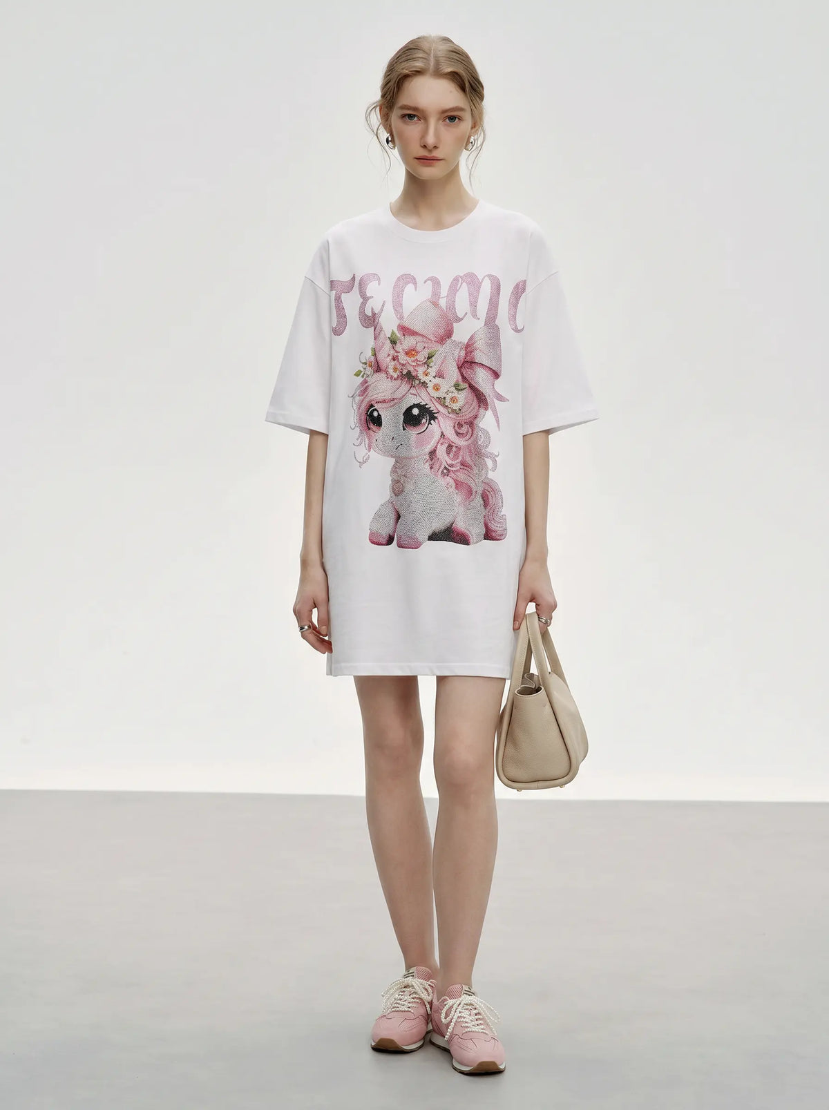 A model stands in a studio wearing an oversized white graphic T-shirt dress with a pink floral illustration and the word 'dream' in pink script. She is holding a beige handbag and wearing pink sneakers. TCH Women's Pink Angel Graphic Oversized T-Shirt