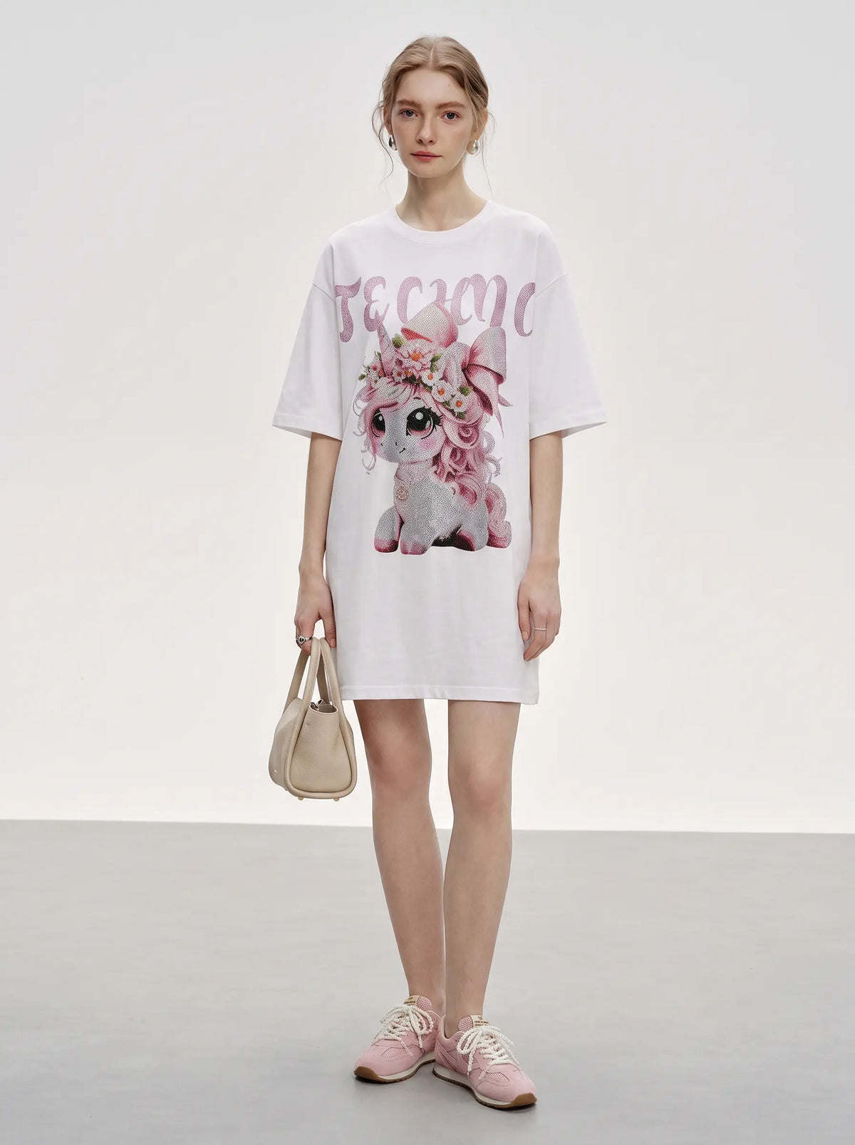 A model stands in a studio wearing a white oversized T-shirt dress with a large pink and white graphic of a flower-adorned dog and the word 'become' across the chest. She is holding a small beige handbag and wearing pink sneakers. TCH Women's Pink Angel Graphic Oversized T-Shirt