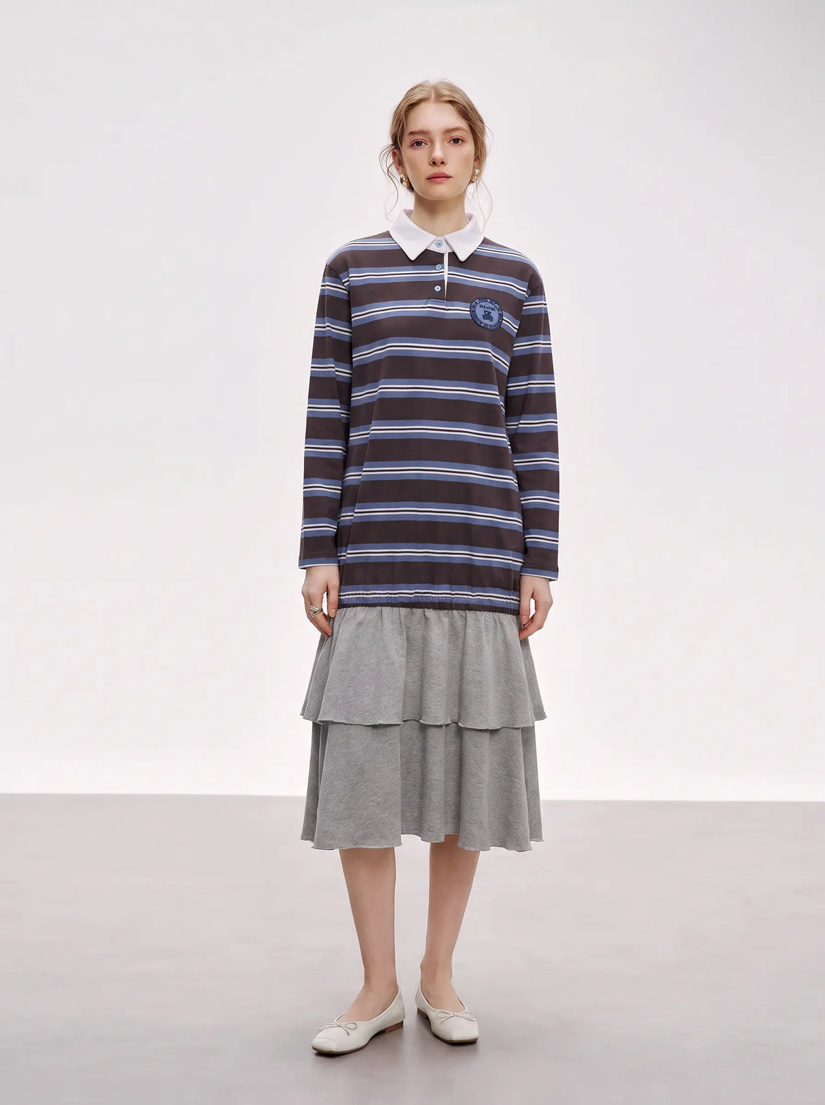 A model stands in a studio wearing a dark navy and blue horizontally striped long-sleeve polo shirt with a white collar, paired with a gray tiered skirt and white pointed flats. TCH Women's Striped Polo Mini Dress