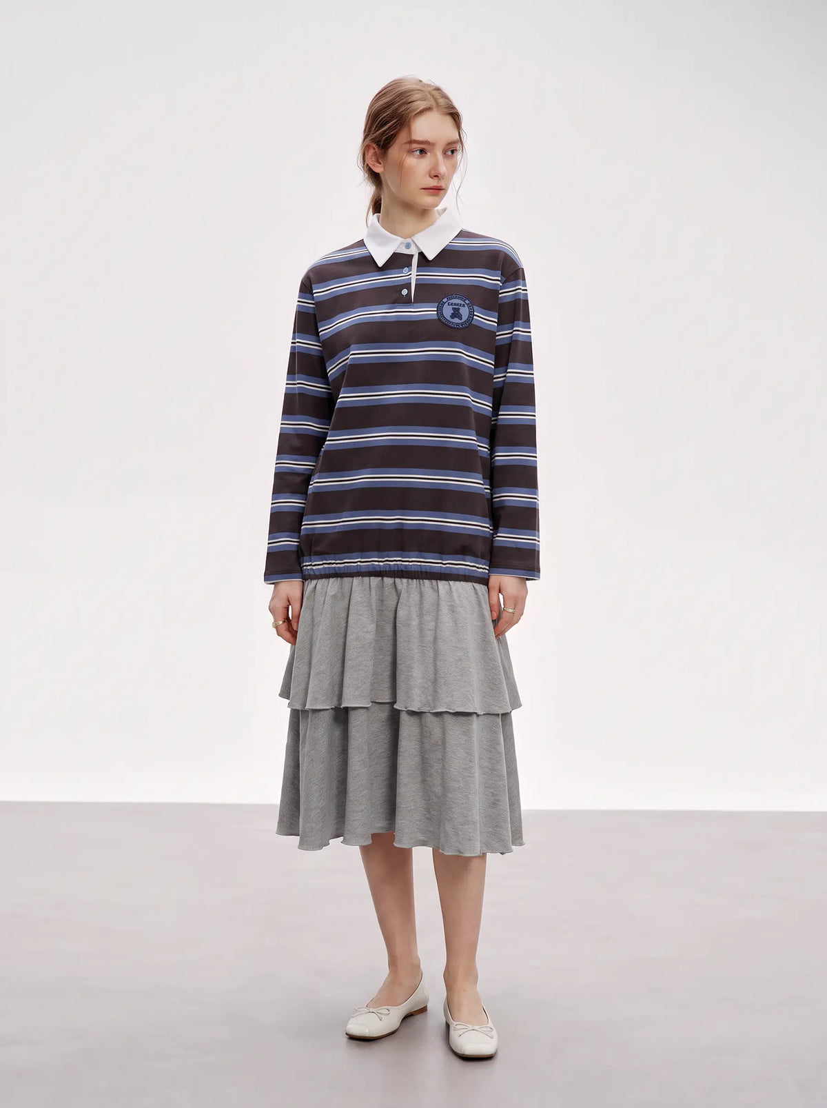 A model stands in a studio wearing a navy and blue striped long-sleeve rugby shirt with a white collar, paired with a gray tiered midi skirt and white flat shoes. TCH Women's Striped Polo Mini Dress