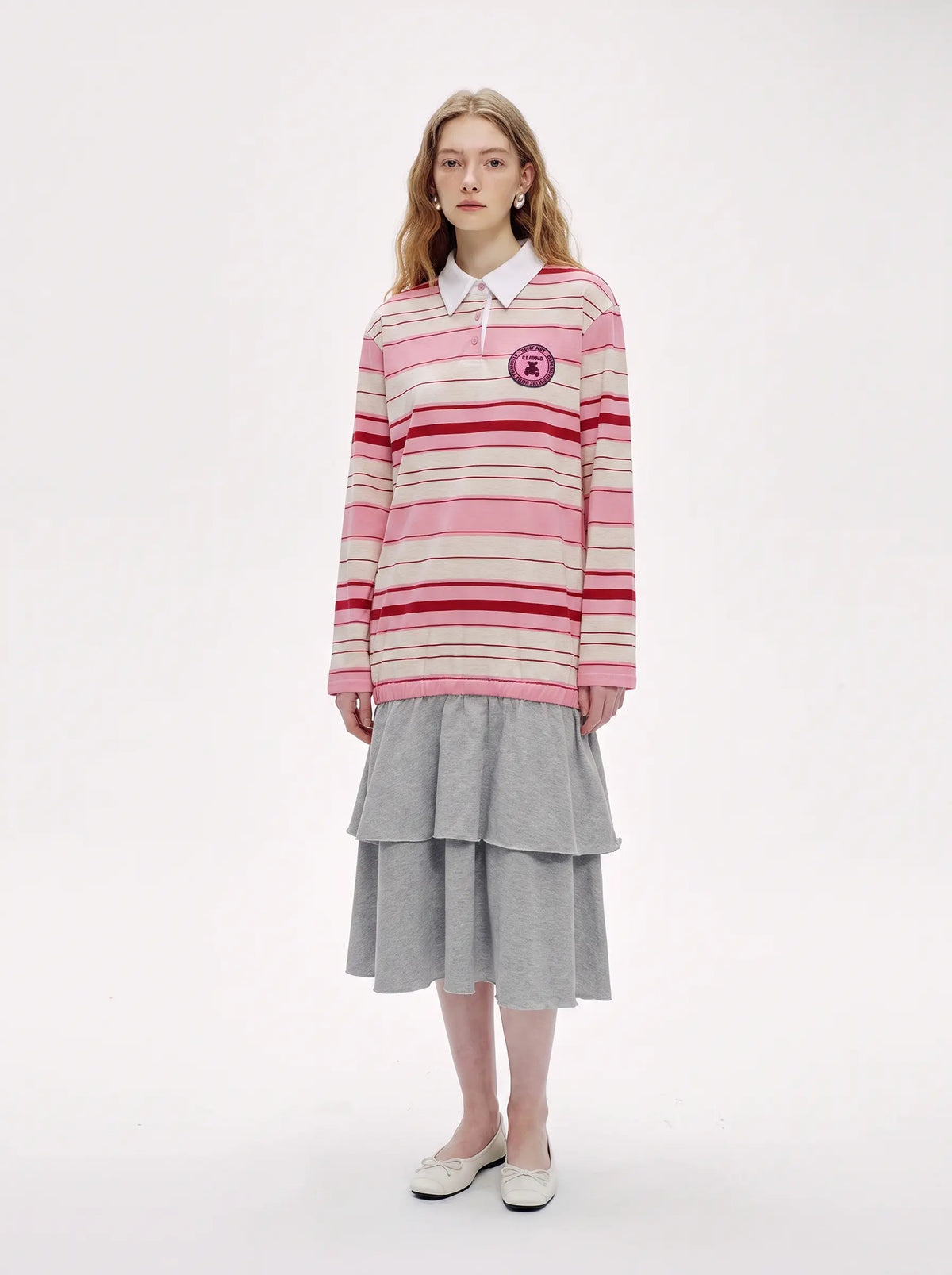 A model stands against a plain white studio background wearing a pink and cream striped long-sleeve polo sweater with a white collar, paired with a gray tiered midi skirt and white shoes. TCH Women's Striped Polo Mini Dress