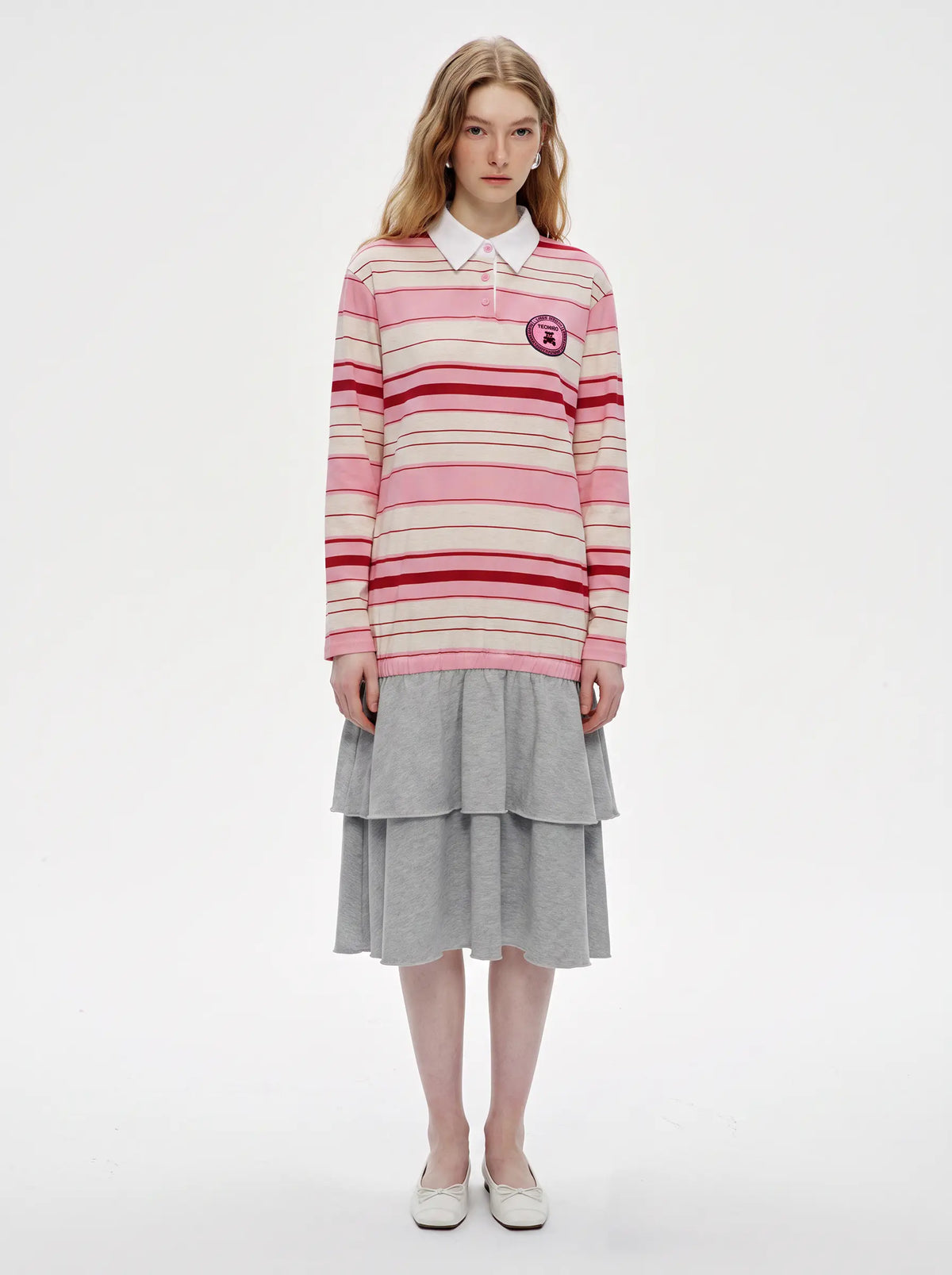 A model stands against a plain white studio background wearing a pink and red striped long-sleeve polo sweater over a white collared shirt, paired with a gray tiered skirt and white shoes. TCH Women's Striped Polo Mini Dress