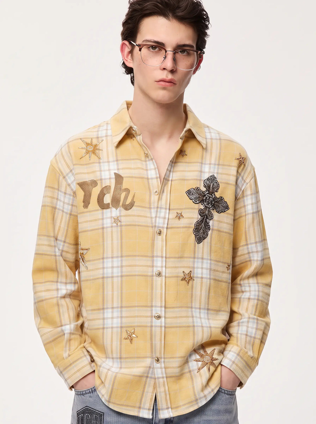 A young man wearing a yellow and white plaid button-up shirt with embroidered details, including a large black rose on the chest, small gold stars, and the letters 'rch' embroidered on the left side. He is also wearing glasses and blue jeans, standing against a plain off-white studio background. TCH Yellow Plaid Embroidered Cross Shirt