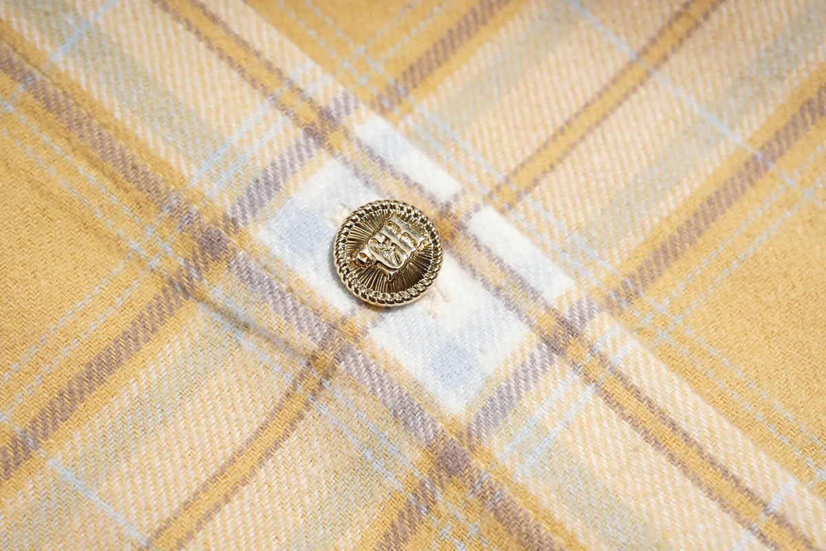 A close-up of a yellow and white plaid fabric with a small round gold button featuring an embroidered monogram. TCH Yellow Plaid Embroidered Cross Shirt