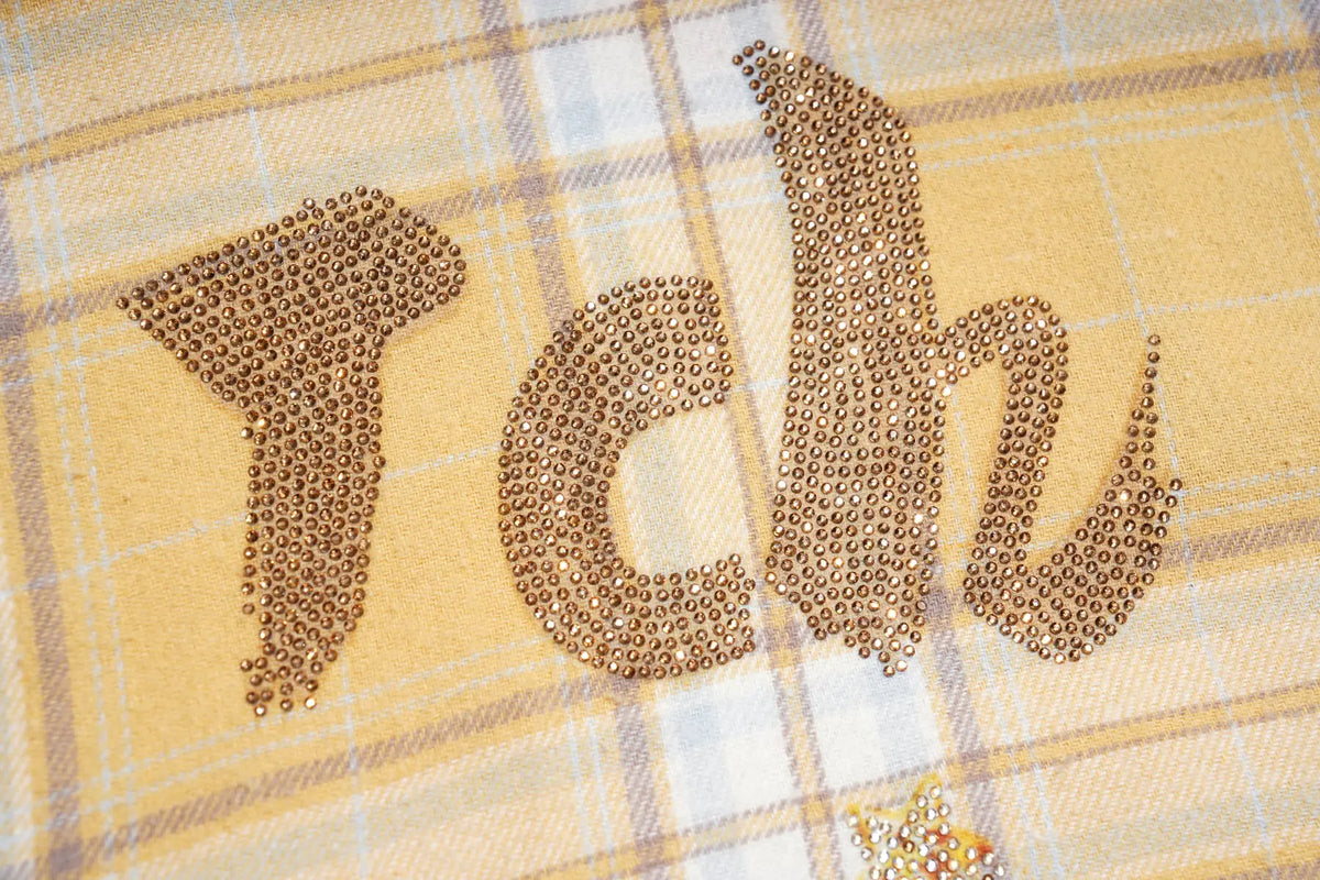 A close-up of a yellow plaid fabric surface with the letters 'Tch' formed from small metallic rhinestones or beads. TCH Yellow Plaid Embroidered Cross Shirt
