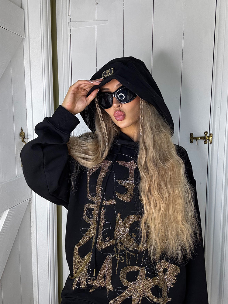 Person wearing a black hoodie with gold designs and a black beanie, standing in a room with white walls and gold door handles.