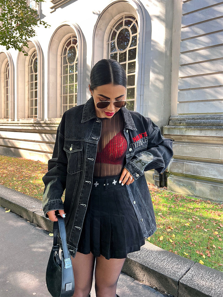 Woman in a red top and black jacket standing outdoors near a building with arched windows.