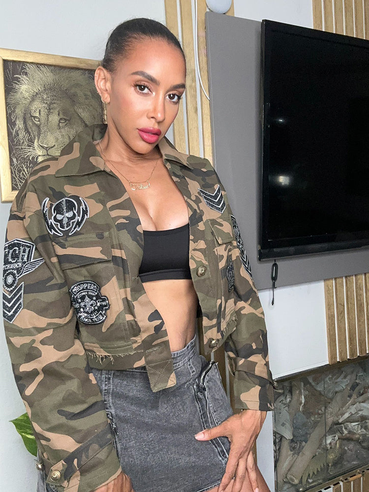 A woman wearing a camouflage cropped jacket with 'TCH' and skull emblems, paired with a black crop top and blue denim shorts, standing in a room with a TV and decorative wall art in the background.