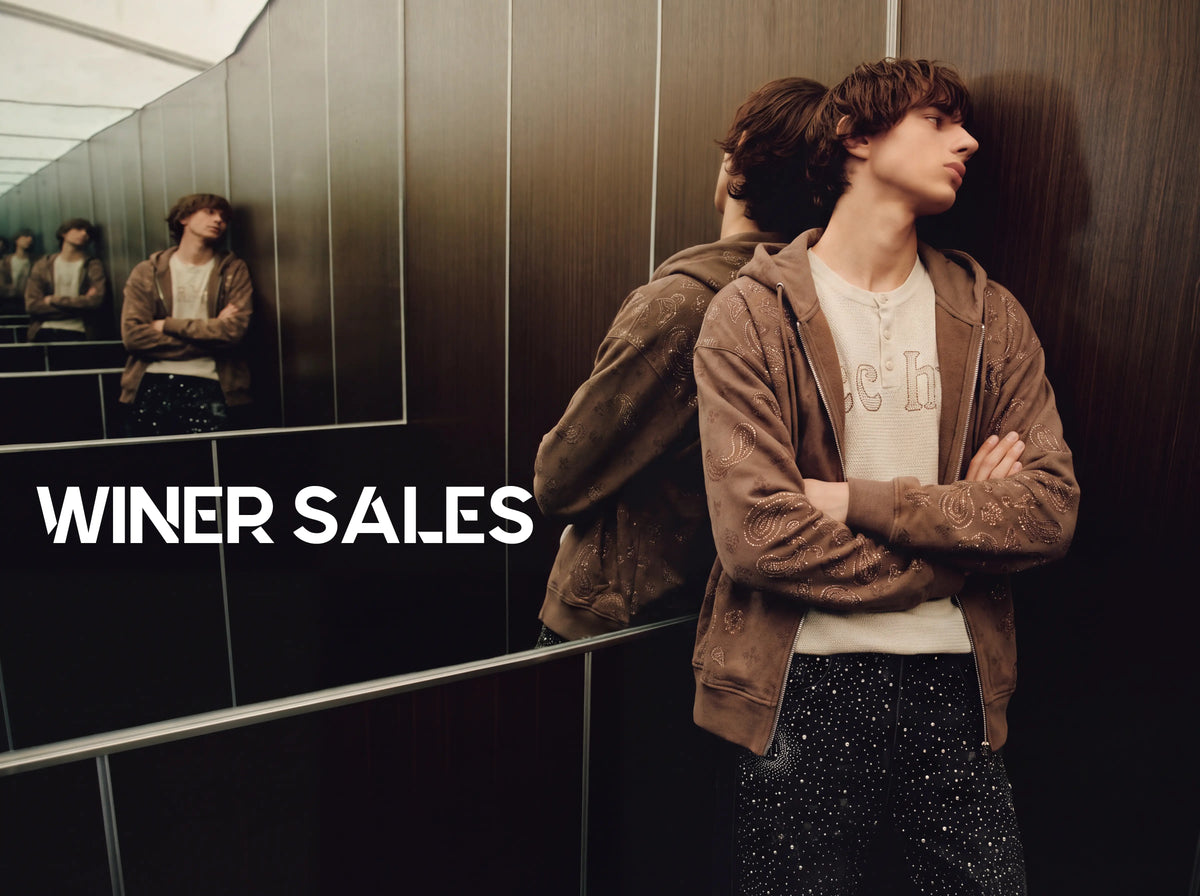 A man stands with arms crossed in a dark interior, wearing a brown zip-up hoodie over a light graphic T-shirt and dark glittery pants. A mirror reflects his back and side view, and large white text on the left reads 'WINER SALES'.