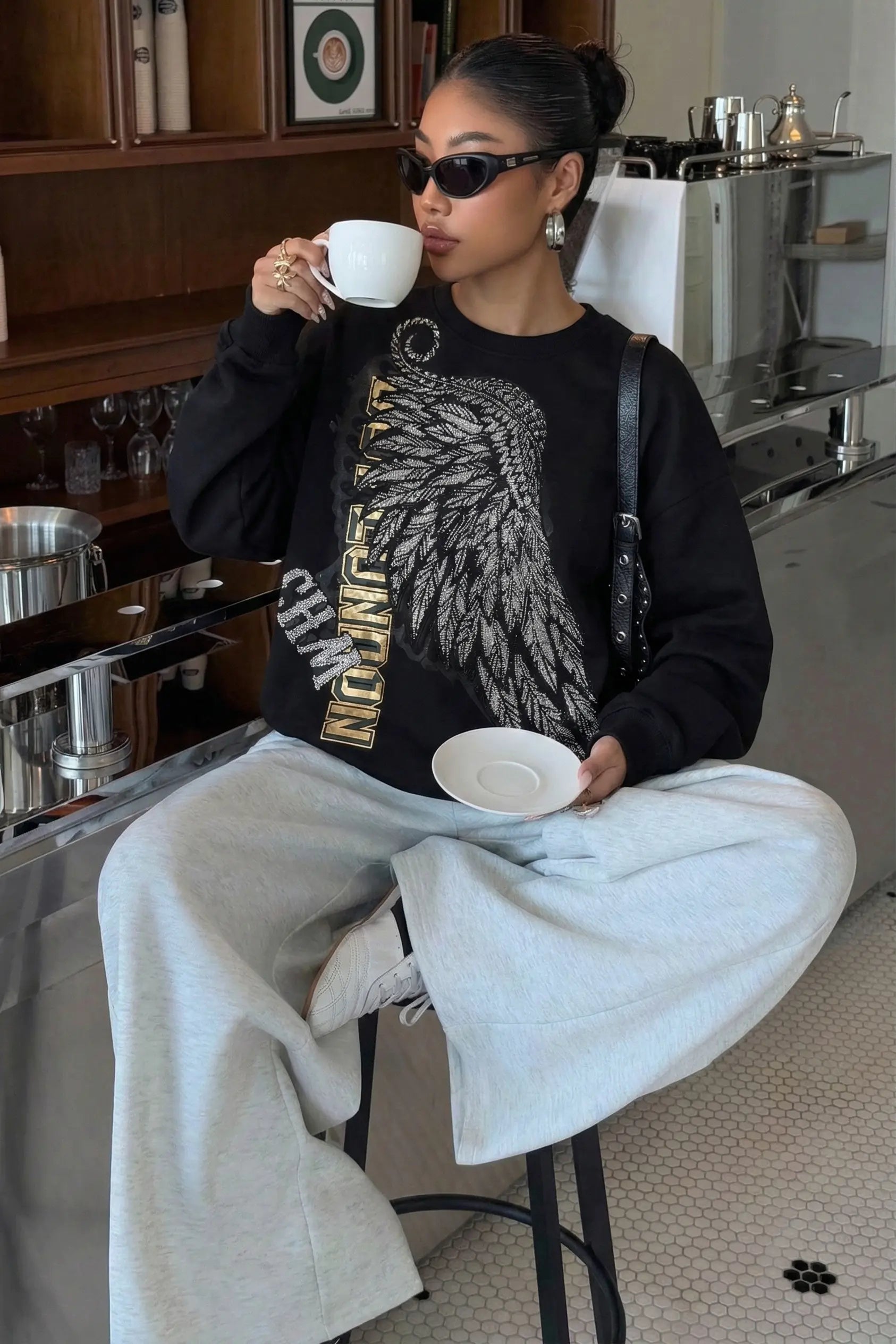 Person sitting in a kitchen holding a cup, wearing sunglasses and a black sweatshirt with a graphic design. Rhinestone Wing Embroidered Sweatshirt