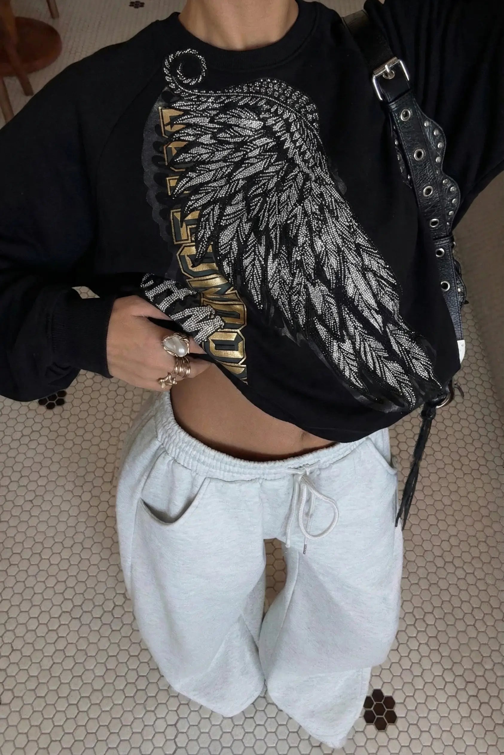 Person wearing a black sweatshirt with a wing design and white pants on a tiled floor. Rhinestone Wing Embroidered Sweatshirt